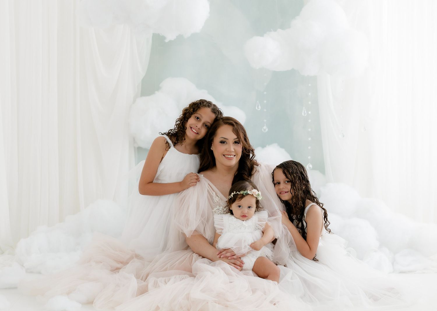A mother in a white gown poses with three daughters in a dreamy white studio with cloud decorations.