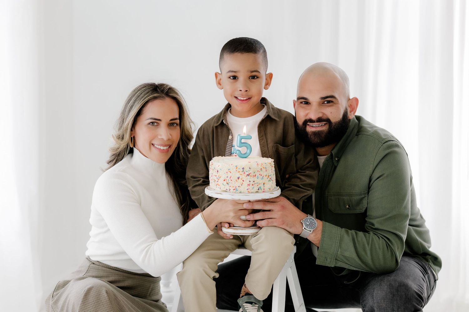 A family poses together holding a birthday cake with the number 5 on top against a white background.