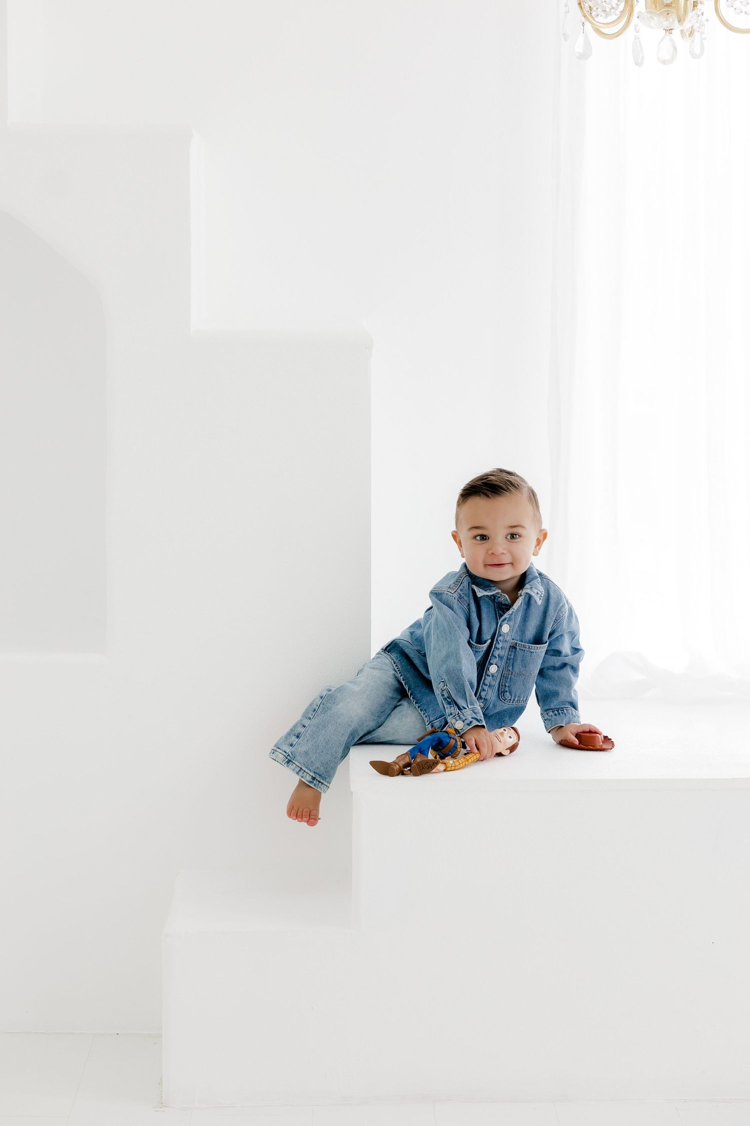 A baby in denim crawls and plays on white blocks in a minimalist studio setting.