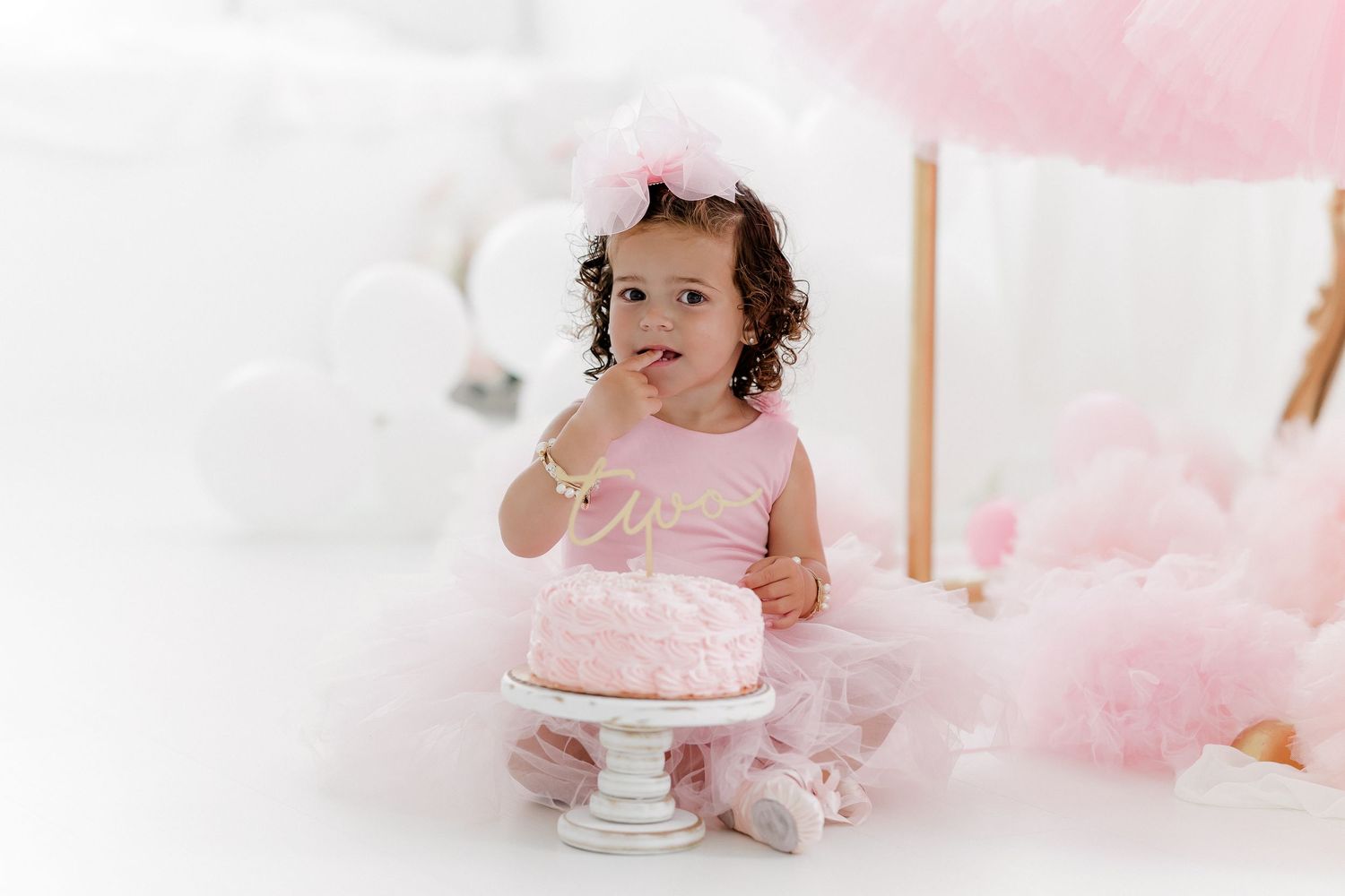 A small child sits next to a pink cake on a white pedestal surrounded by soft pink tulle decorations.