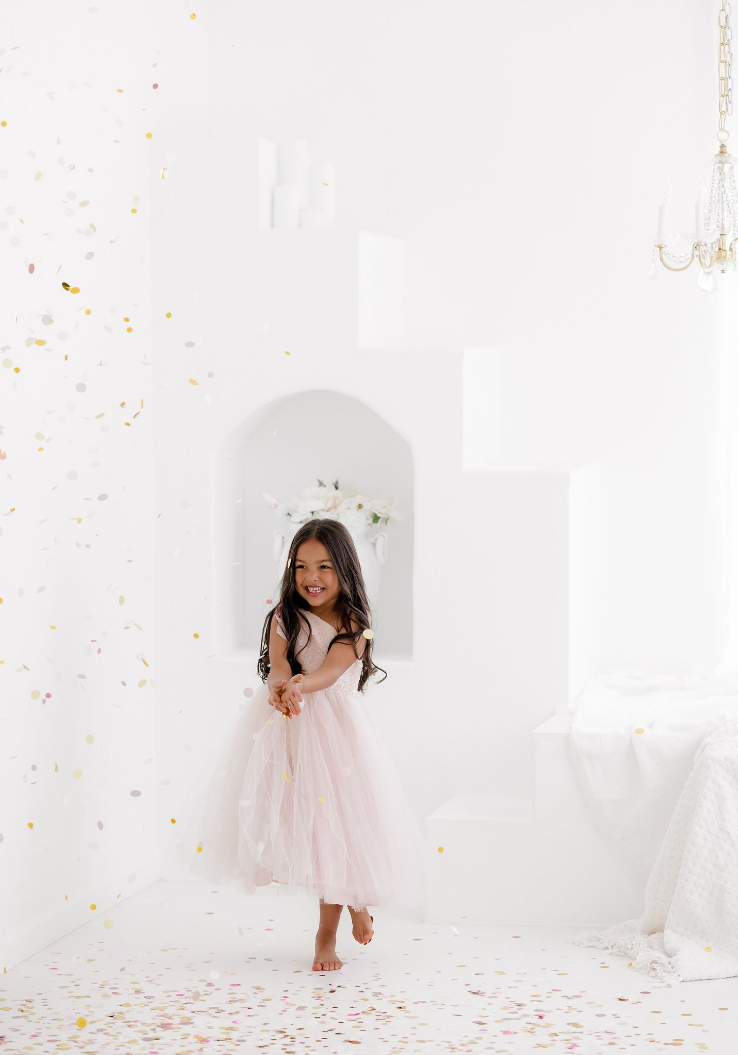 A young girl in a pink tulle dress twirls and plays with confetti in a bright white studio setting.