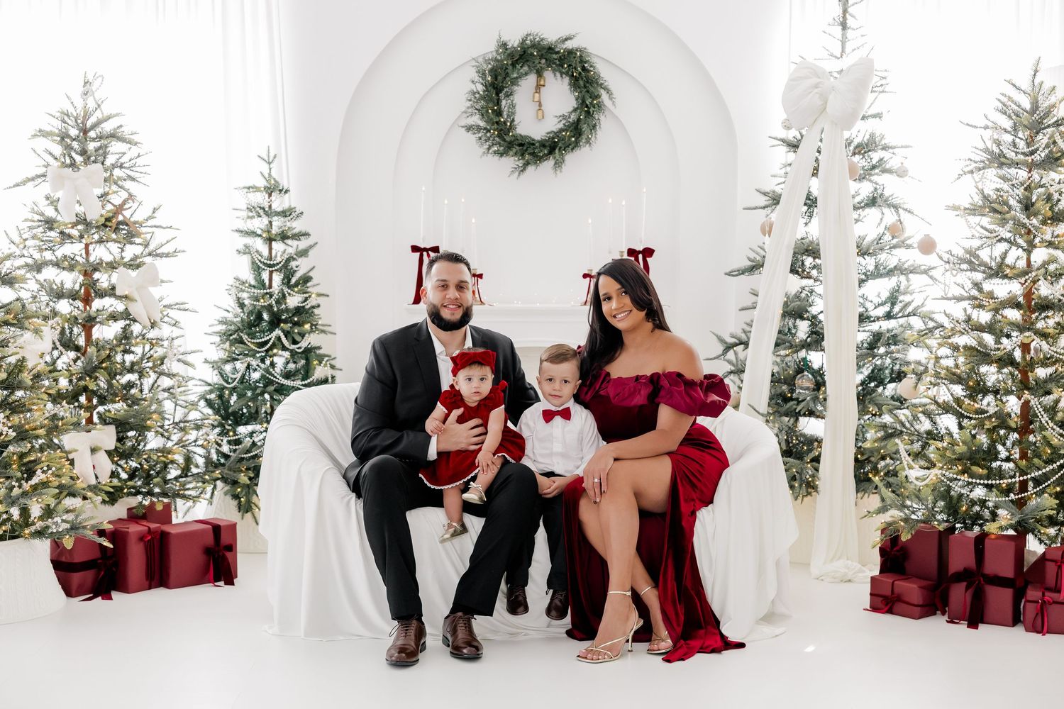 Family of three in red and black holiday outfits posing with Christmas trees and gifts in a white studio.