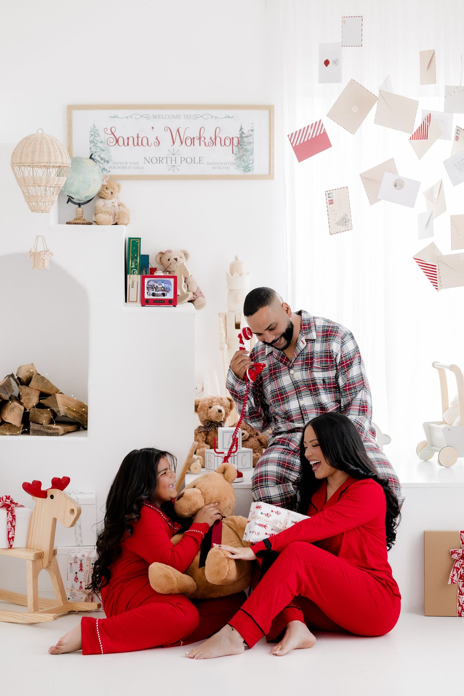 Family in red and plaid pajamas celebrating Christmas morning with teddy bears and gifts in a Santa's Workshop decorated room.