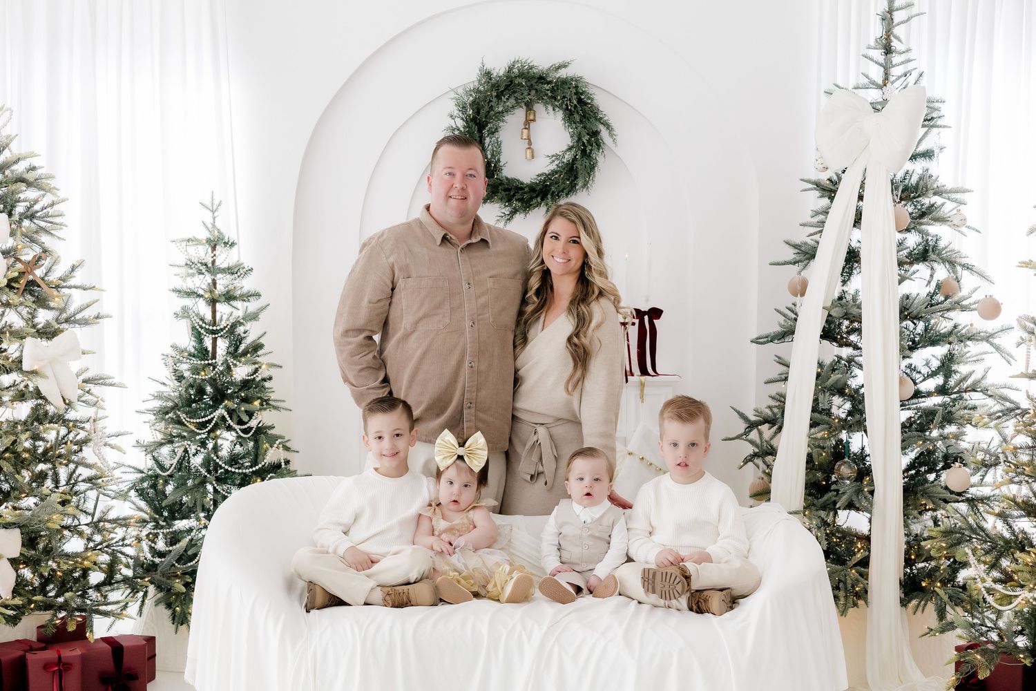Family of five poses with Christmas trees and wreath in bright white studio for festive holiday portrait.