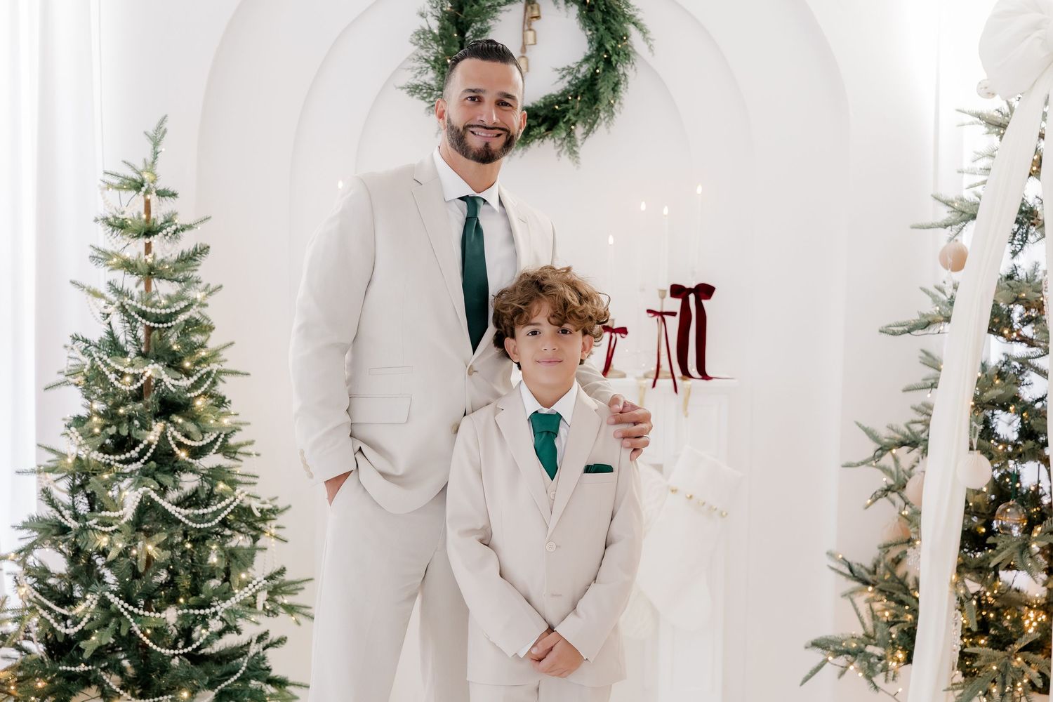 Man and boy in matching white suits pose by Christmas trees with holiday decor and wreath backdrop.