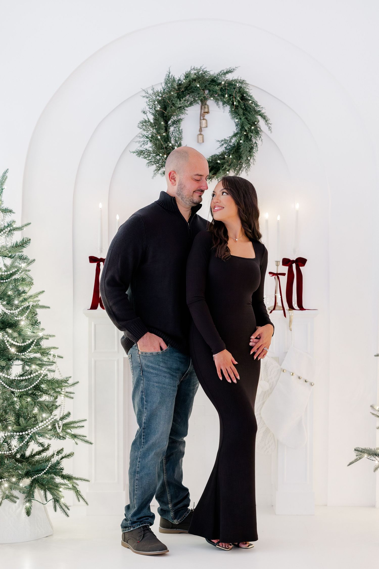 Couple in black outfits posing by Christmas tree and wreath decor in white studio for holiday maternity photo session.