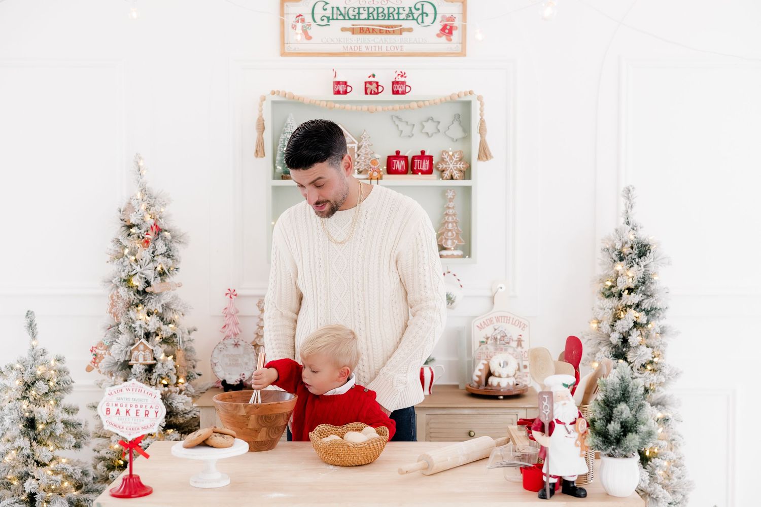 Father and toddler baking Christmas cookies in a festive white kitchen with decorated trees and gingerbread decor.