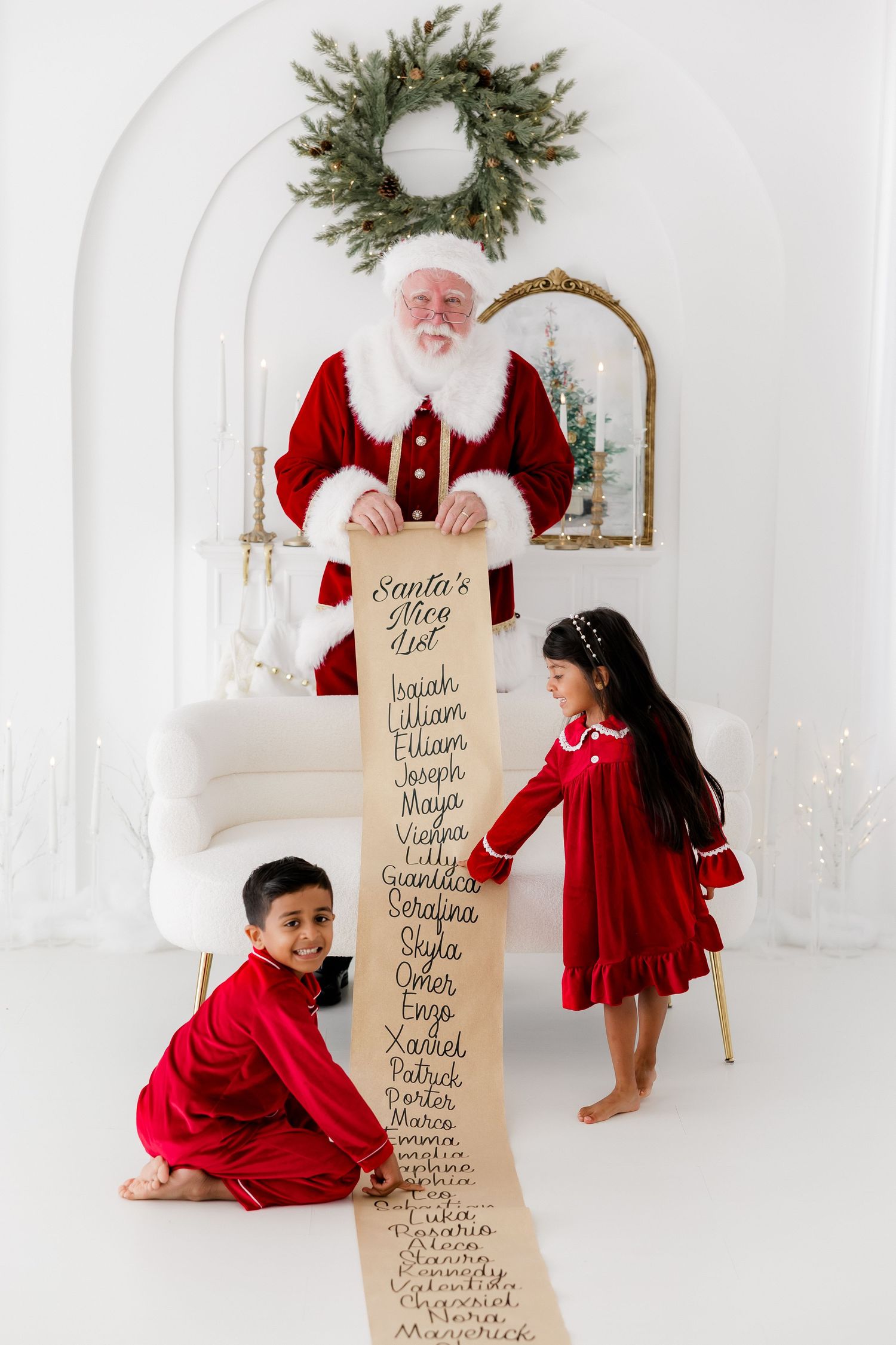 Santa holds his Nice List scroll with two children in red outfits in a white Christmas studio.