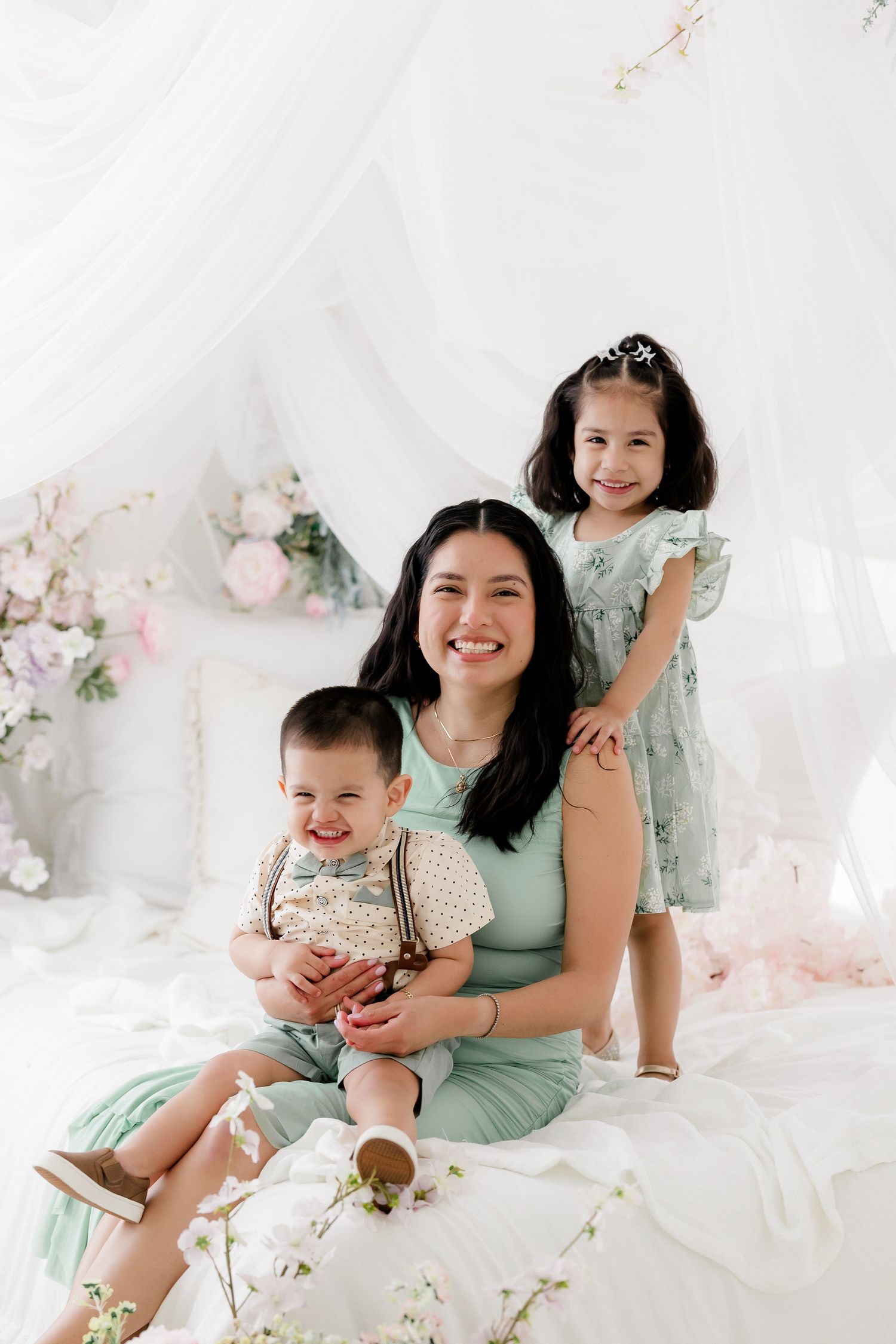 Family wearing mint green dresses poses together on white bedding with floral decorations in studio setting.