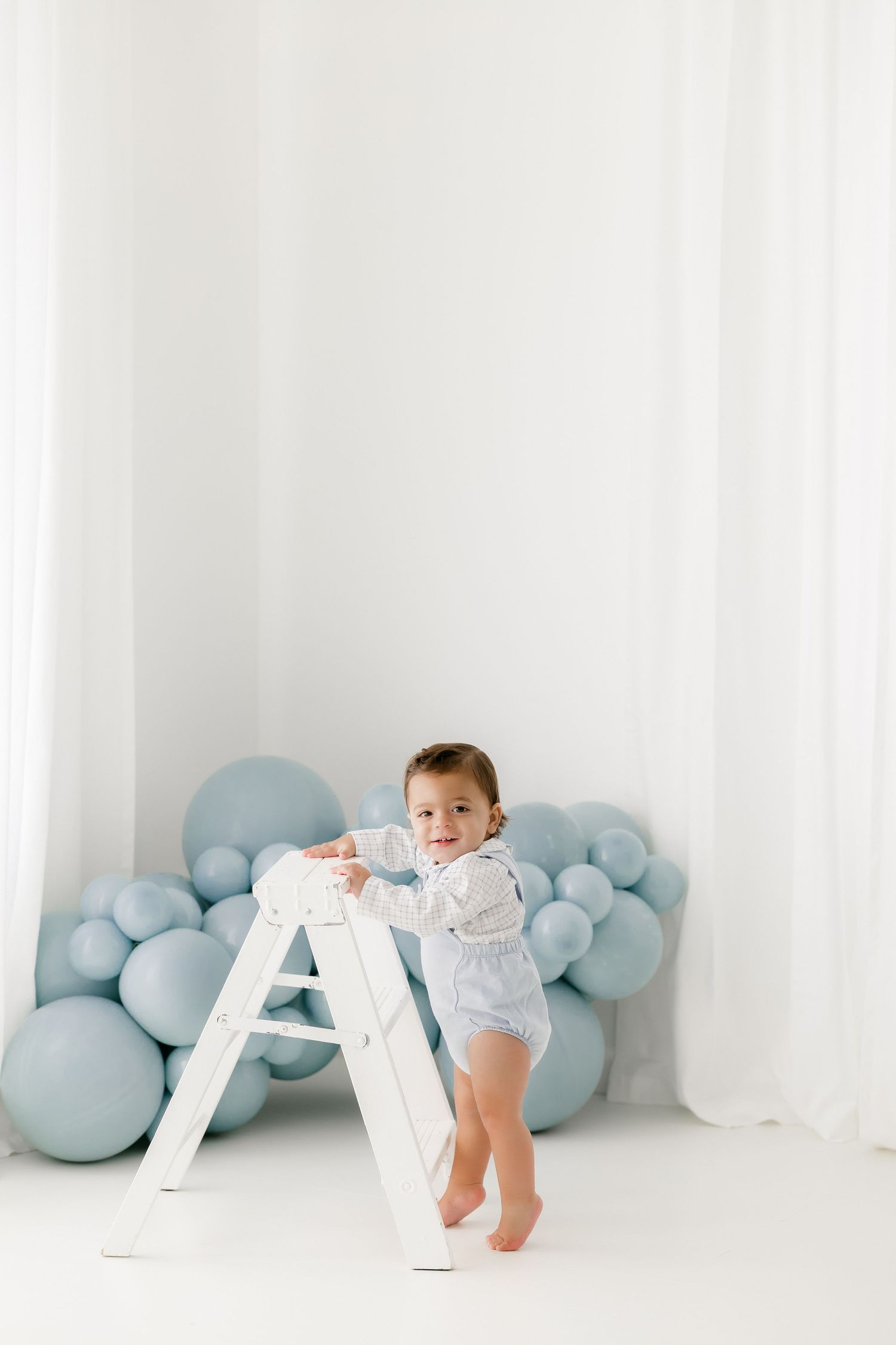 A baby in white clothing crawls on a white floor with soft blue balloon decorations in the background.