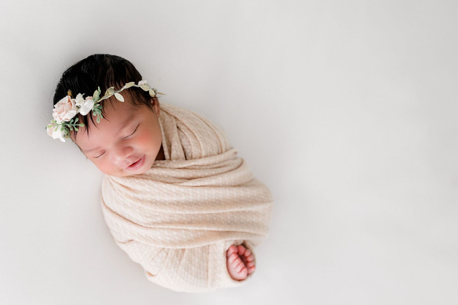 Newborn baby wrapped in beige cloth wearing delicate white flower crown against white background.