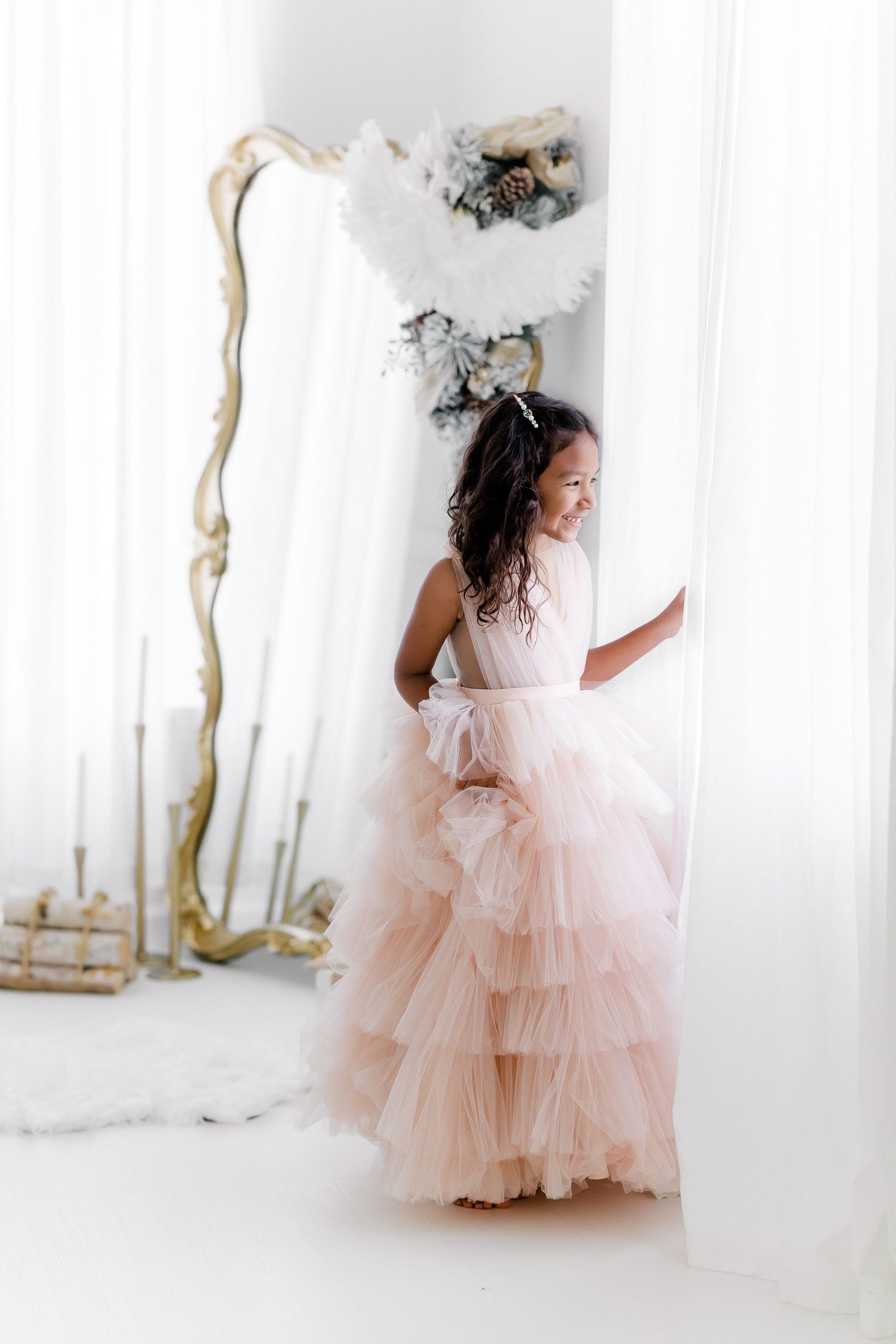 Elegant ruffled pink gown photographed in bright white room with ornate mirror and decor.