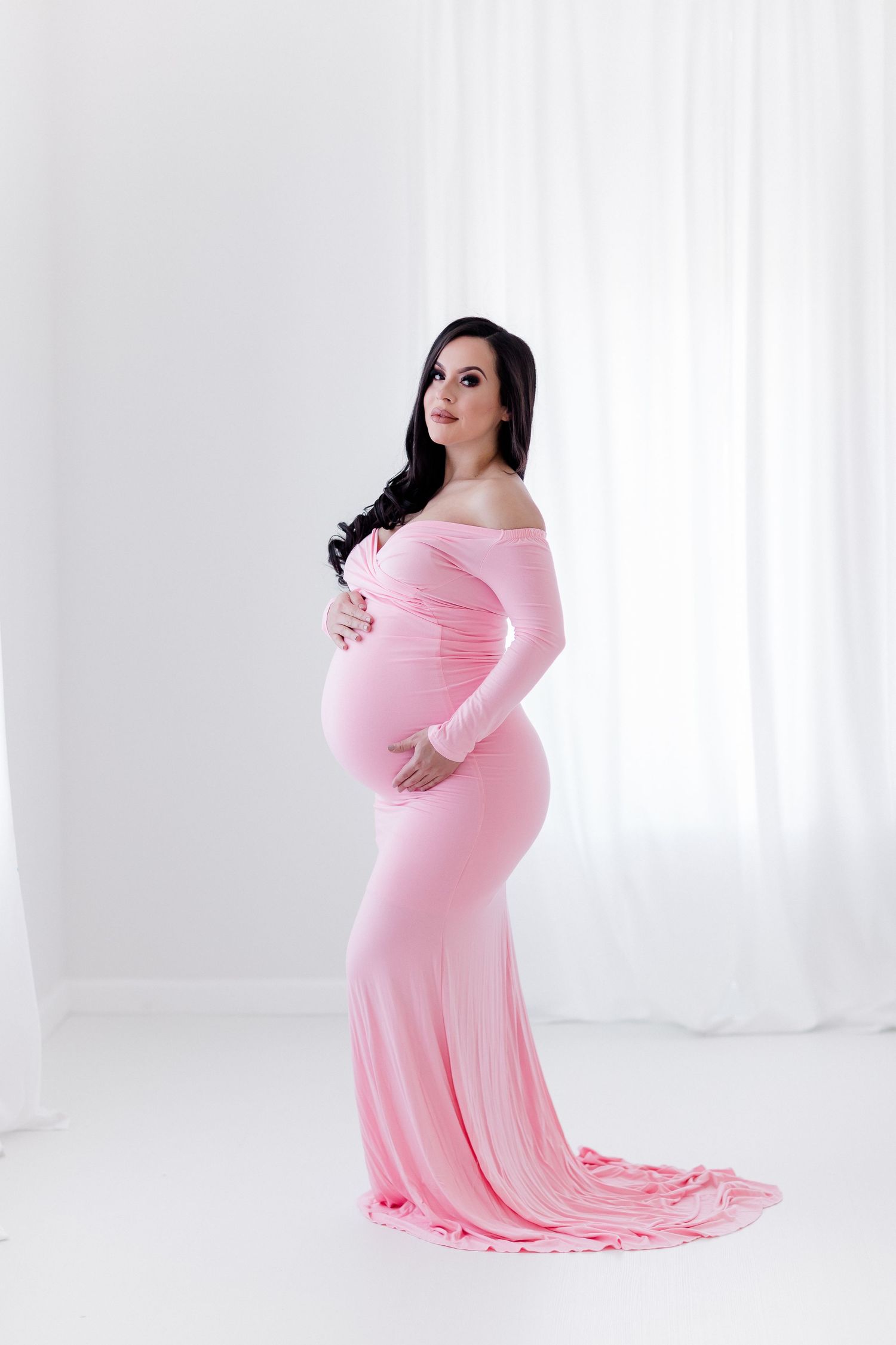 Elegant maternity portrait in form-fitting pink gown against white studio backdrop.