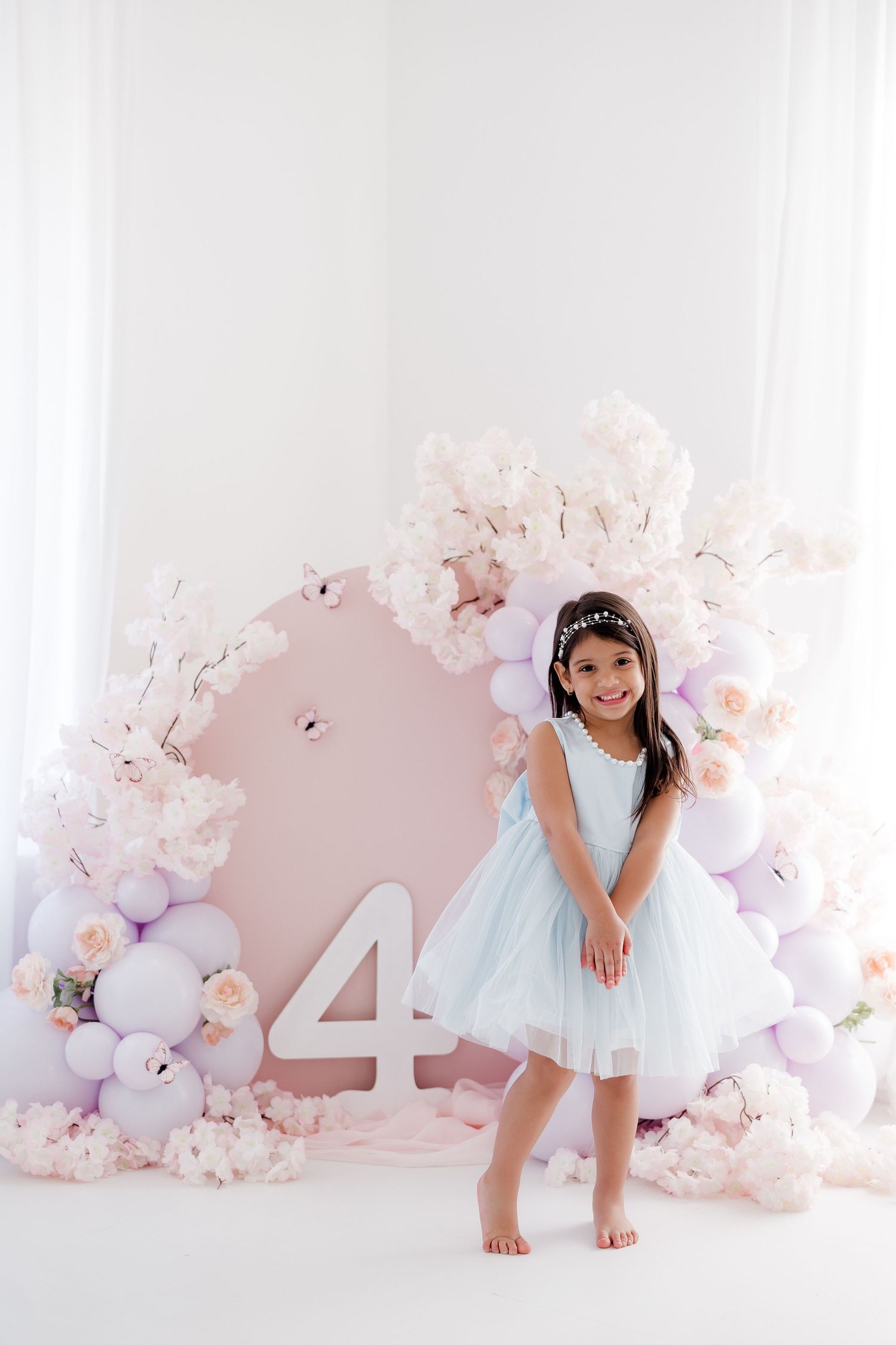 A smiling girl in a blue dress poses by a pink balloon arch with floral decor and a number 4 for her birthday.