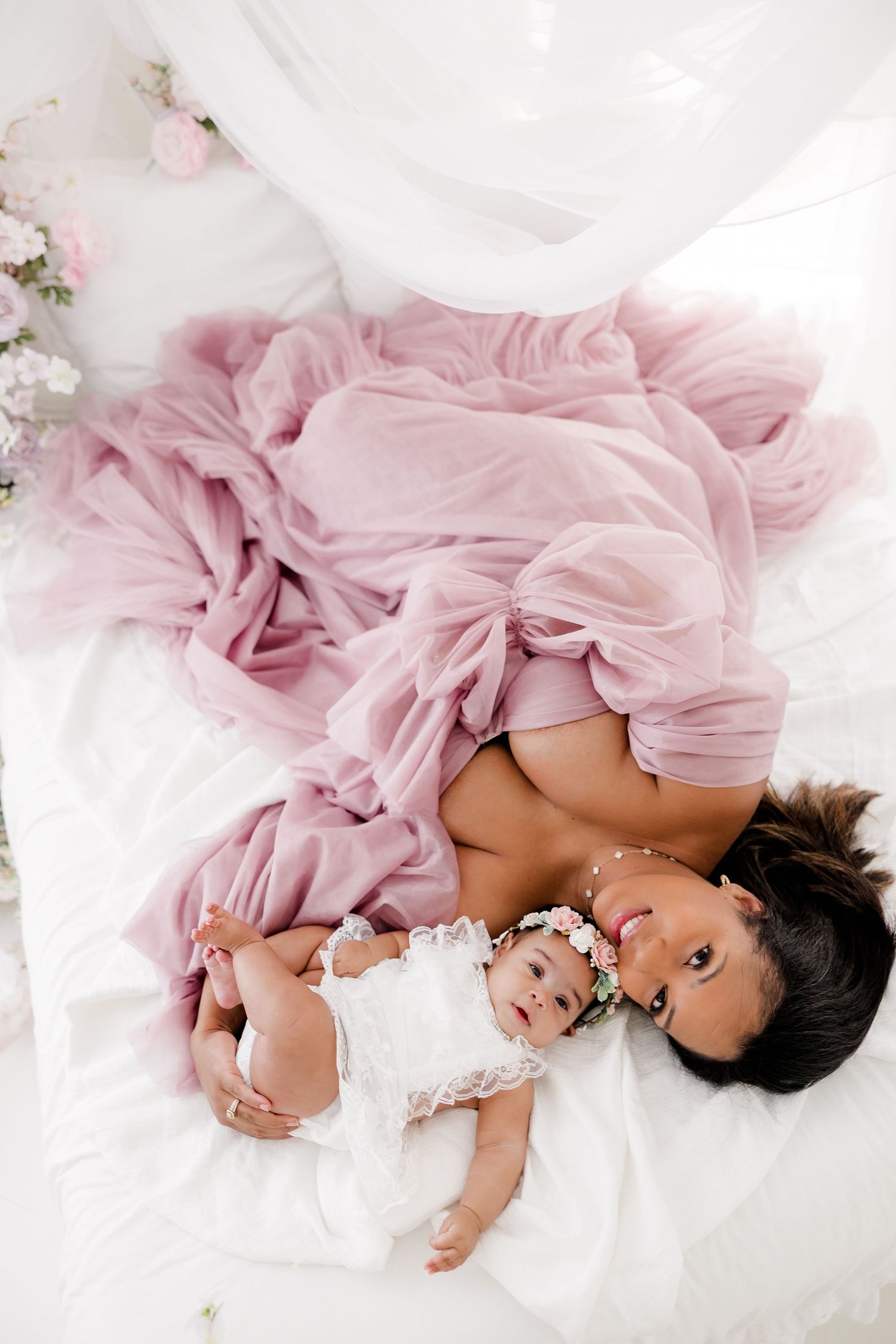 A baby in a flowing pink tulle dress lies on white bedding surrounded by delicate floral accents and soft lighting.