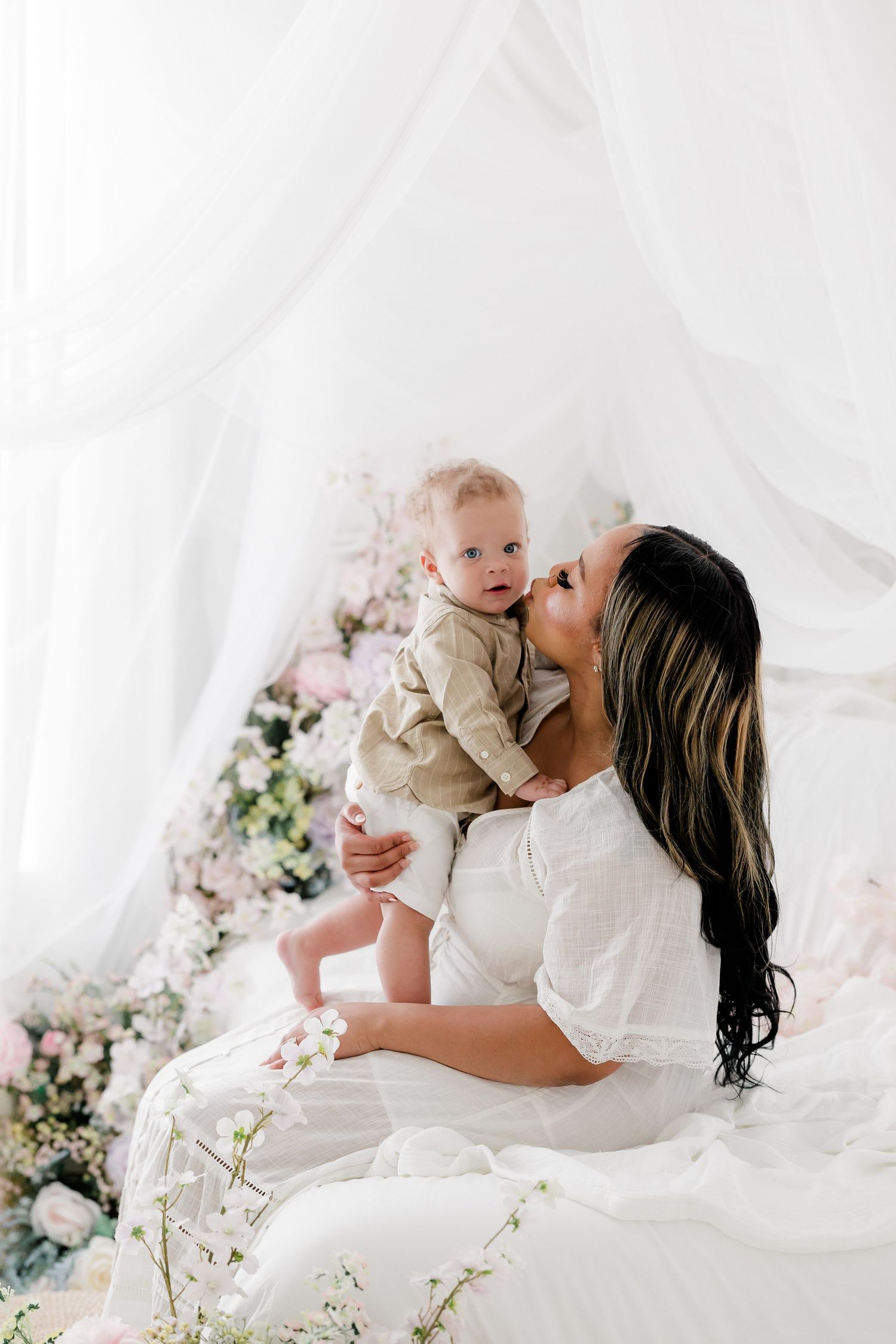 A tender moment between parent and baby surrounded by white fabric and delicate floral decorations in a bright setting.