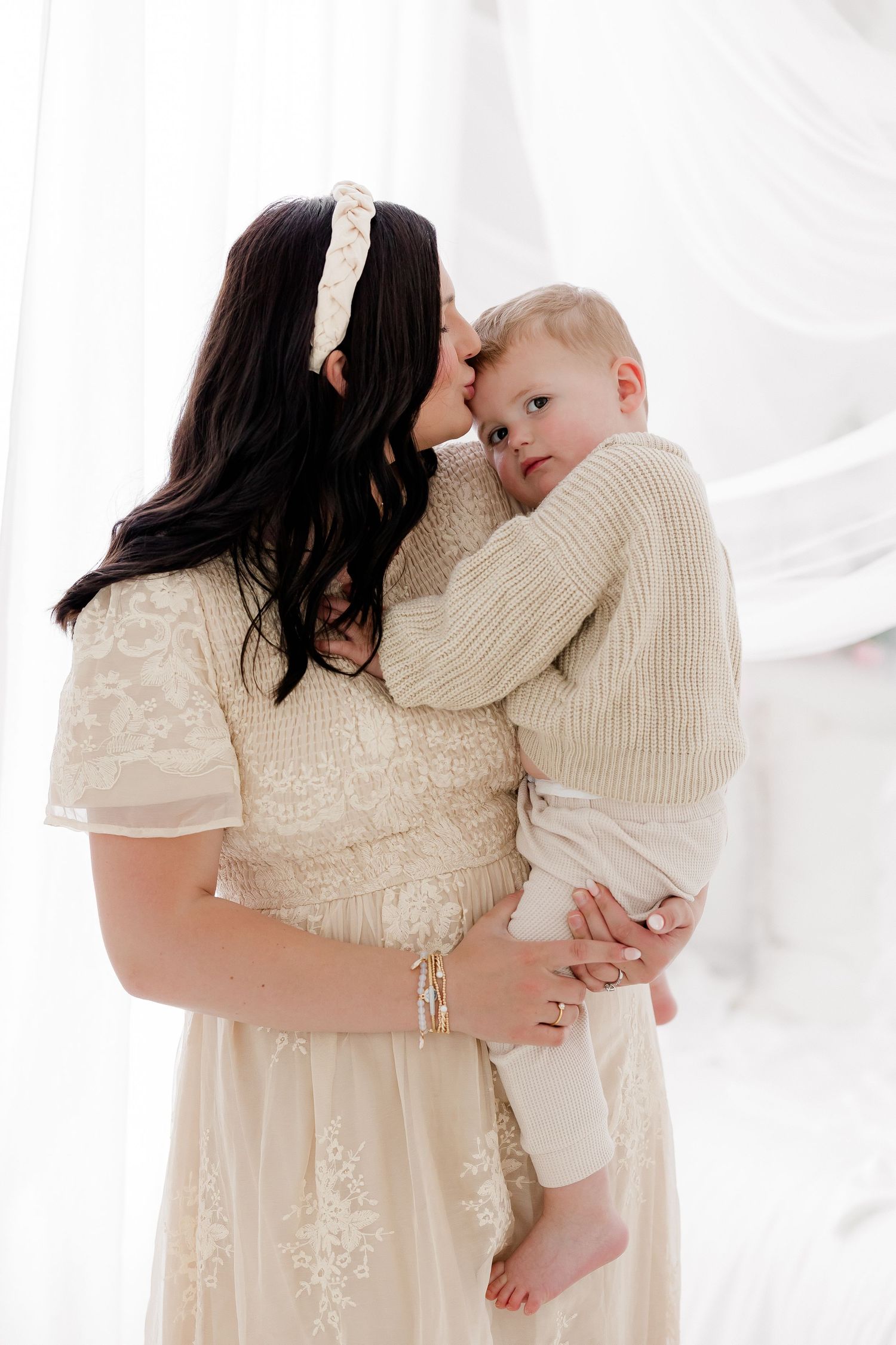 A tender moment between parent and baby in white outfits against a bright white backdrop in a studio setting.