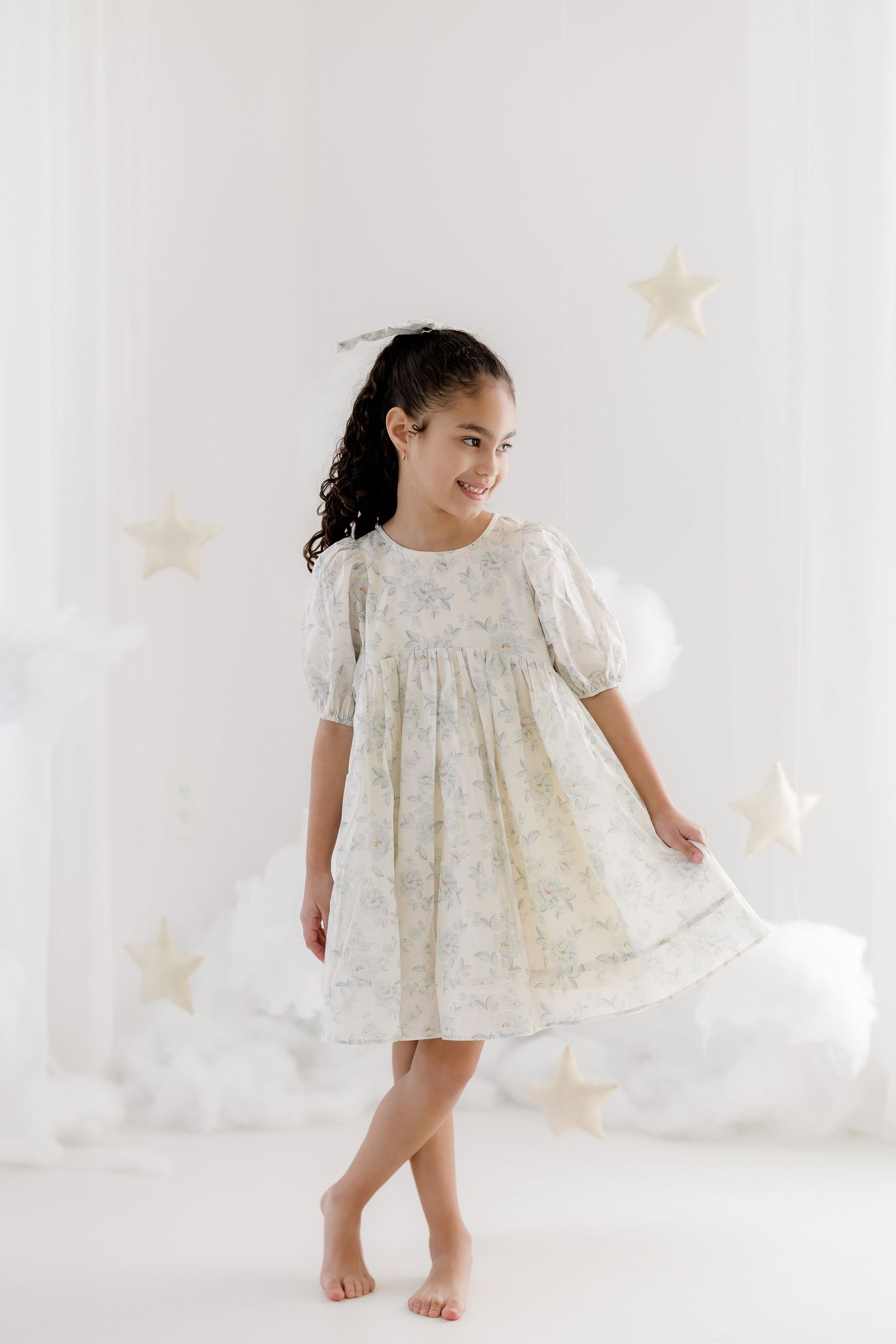Young girl in a floral pastel dress twirling barefoot in a dreamy white studio with star decorations.