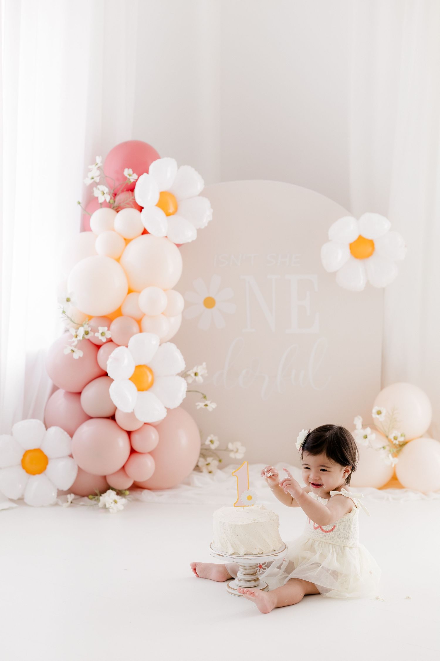 Pastel pink balloon arch with white daisy decorations creates a dreamy backdrop for a baby's cake smash photo session.