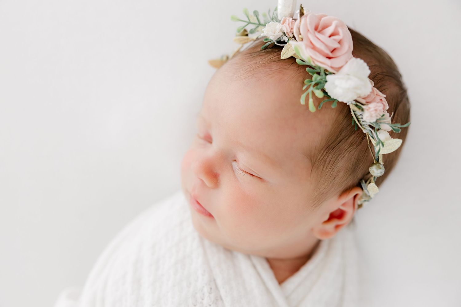 Newborn baby sleeping peacefully wearing delicate pink and white floral crown against white background.