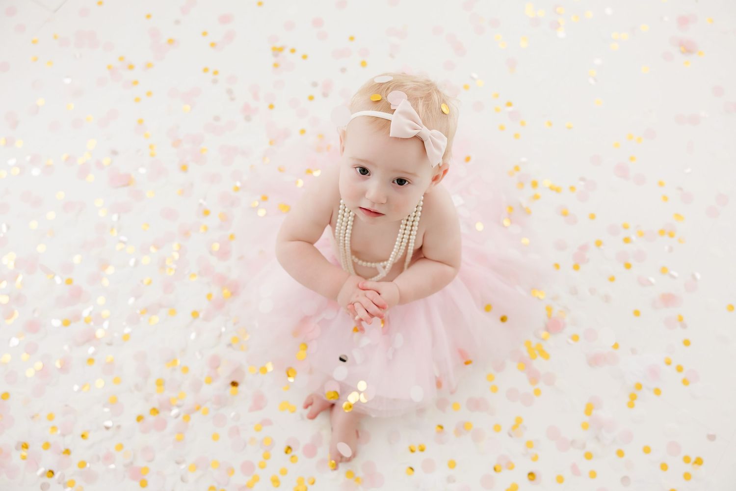 Baby in pink tutu dress crawls on white backdrop surrounded by colorful confetti during first birthday photoshoot.