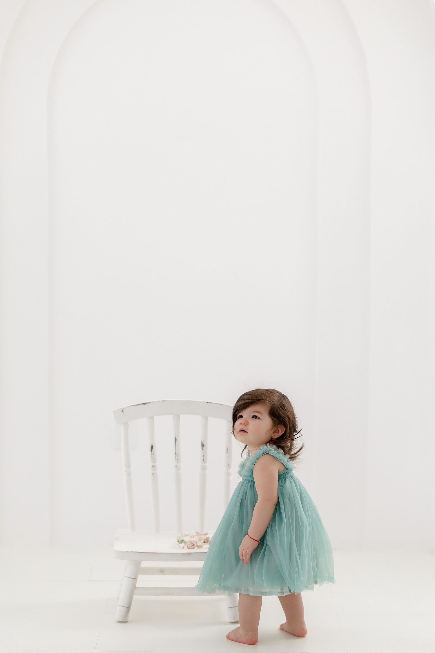 A child in a mint green tutu dress plays and poses around a white wooden chair in a bright studio setting.
