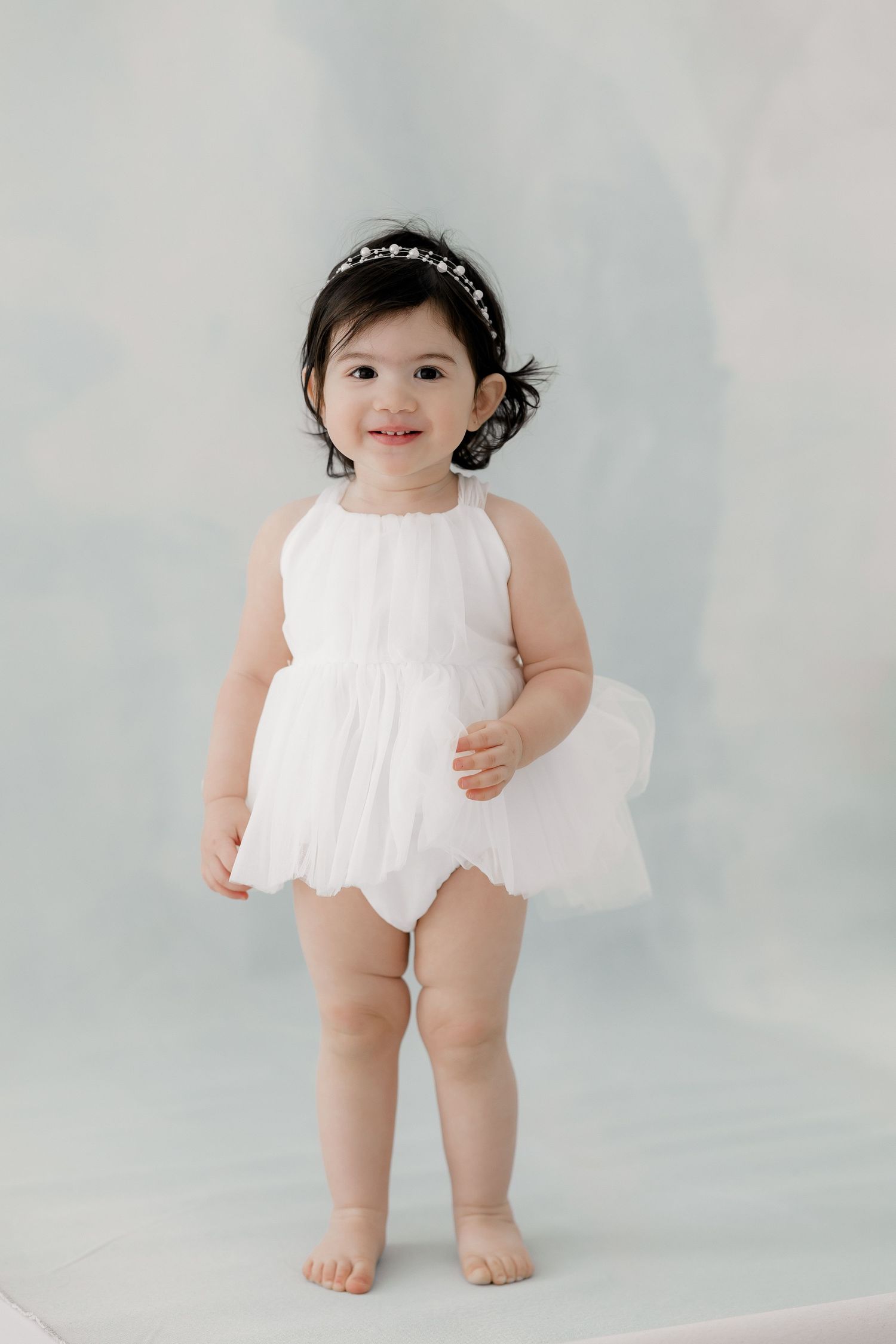 Sweet baby in white lace romper poses against light blue backdrop for professional studio photography session.