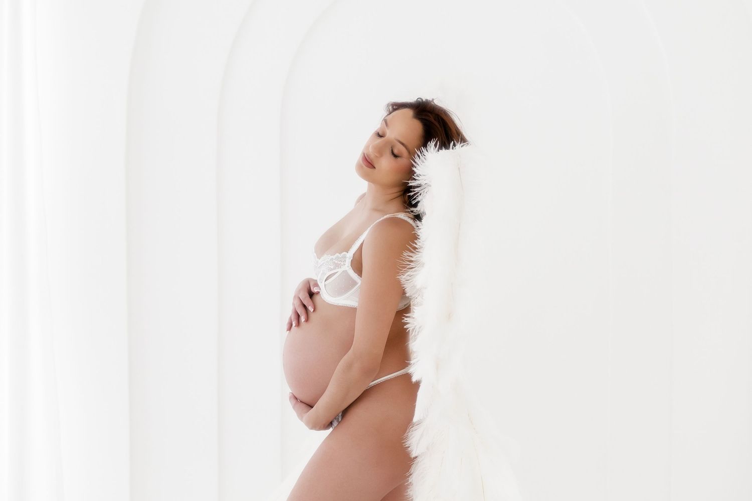A pregnant woman in white lingerie poses against a light background during a maternity photoshoot.