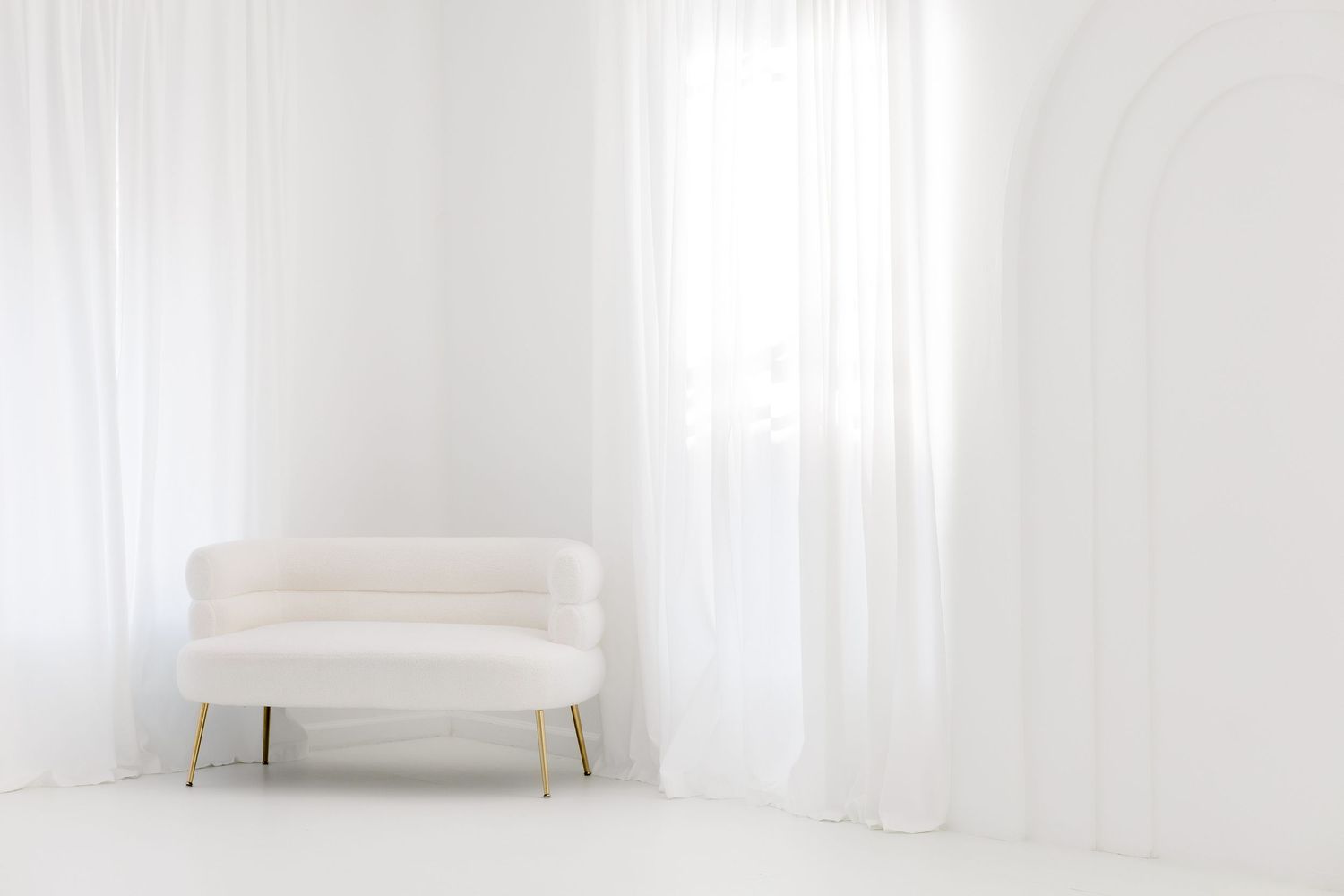 Modern white couch sits elegantly against a bright window with sheer curtains in a minimalist living room setting.