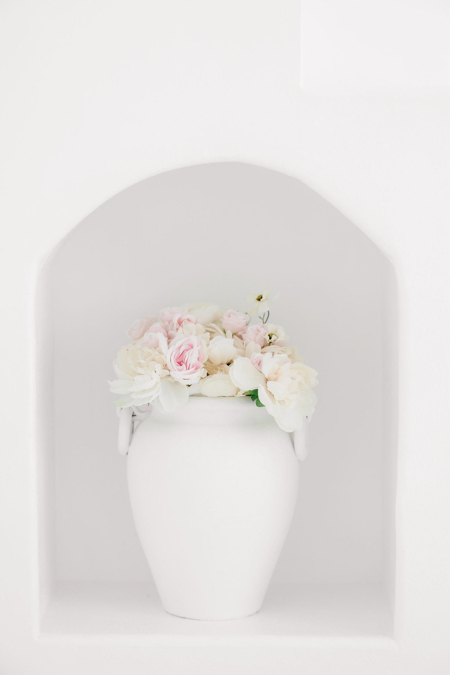 A white ceramic vase with delicate pink and white roses arranged in a minimalist style against a soft gray background.