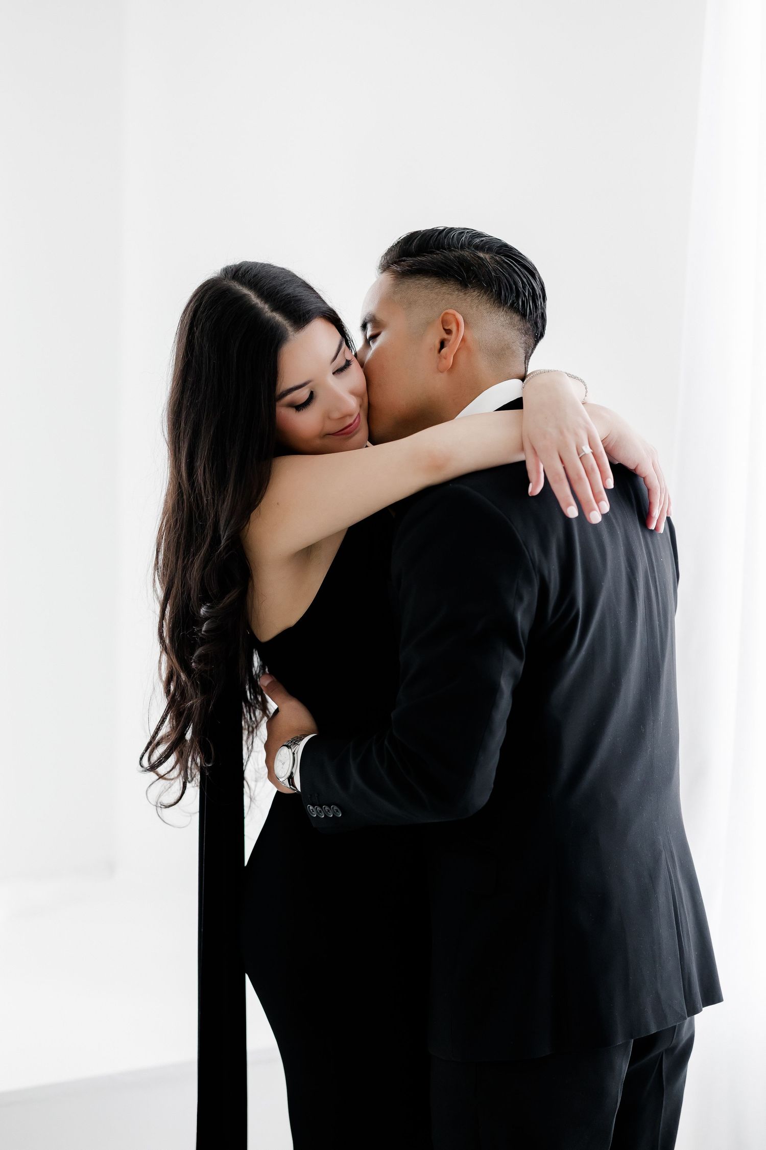 A couple in black attire embraces intimately against a white background in a romantic photoshoot.