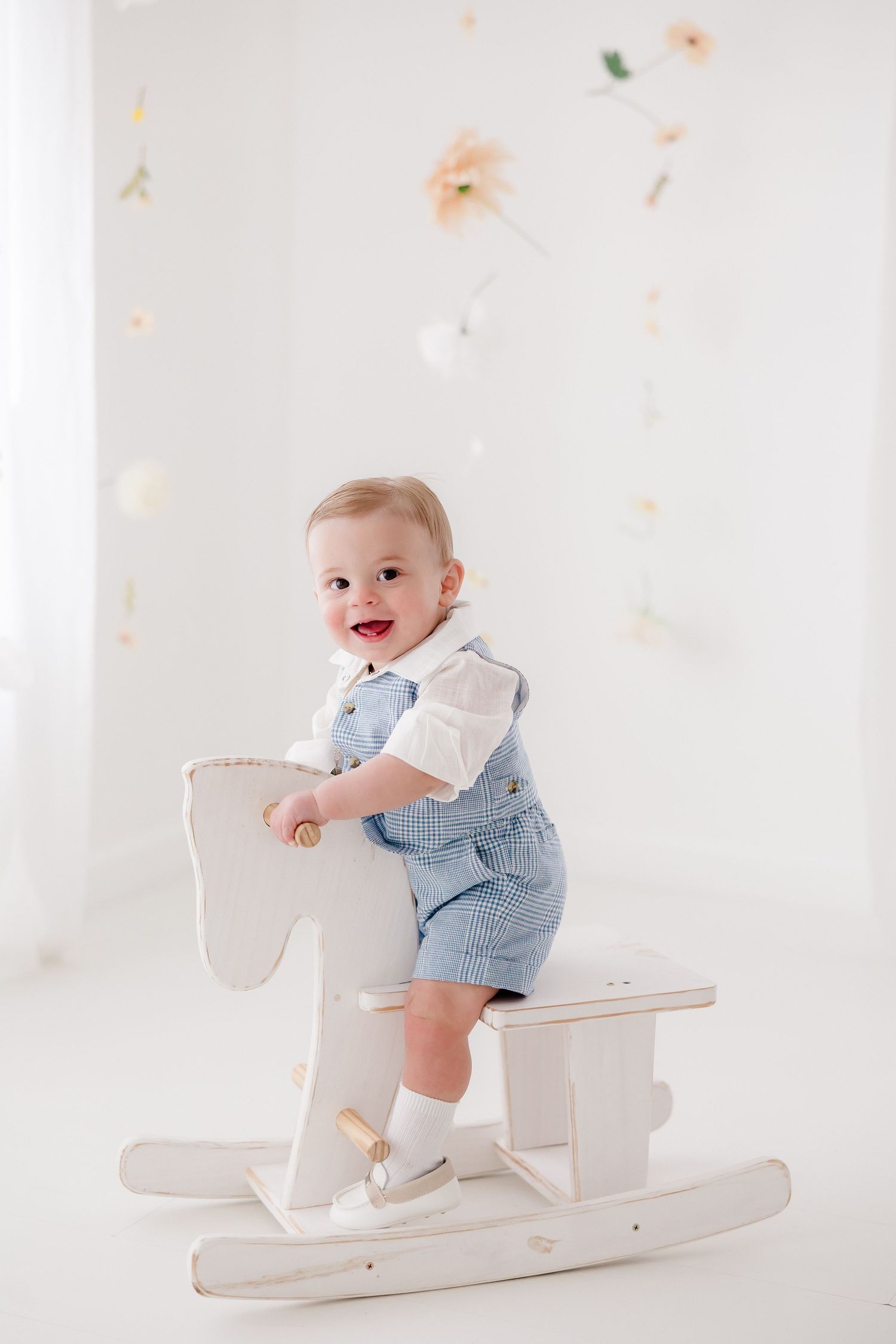 A baby wearing blue overalls sits and plays on a white wooden rocking horse against a light floral backdrop.