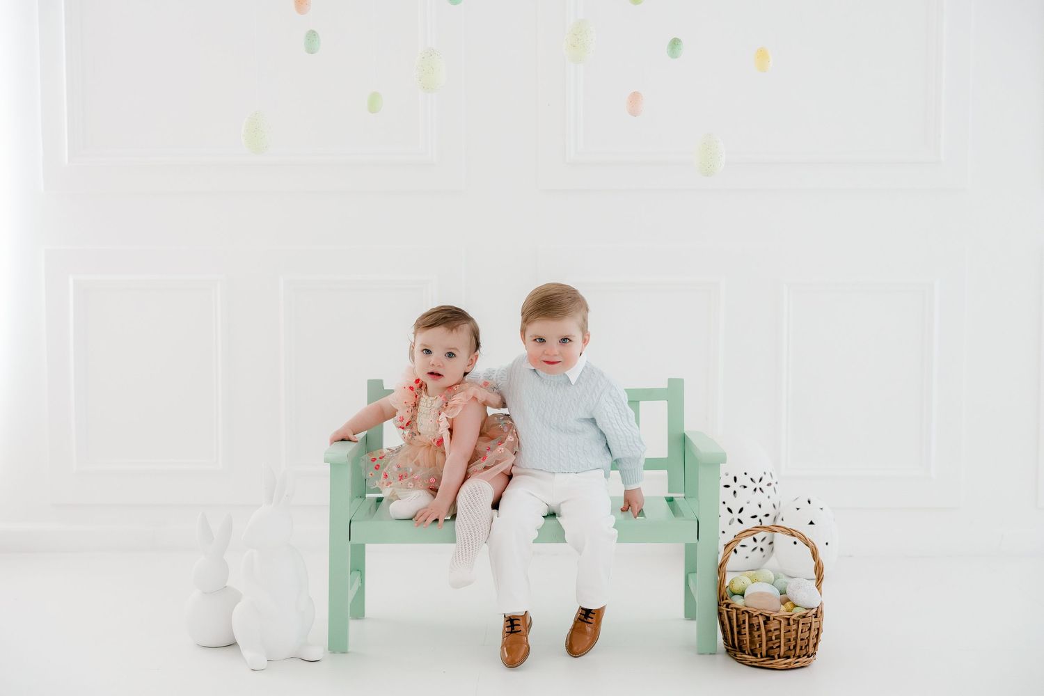 Two children sit on a mint green bench with a woven basket and stuffed animal beside them.