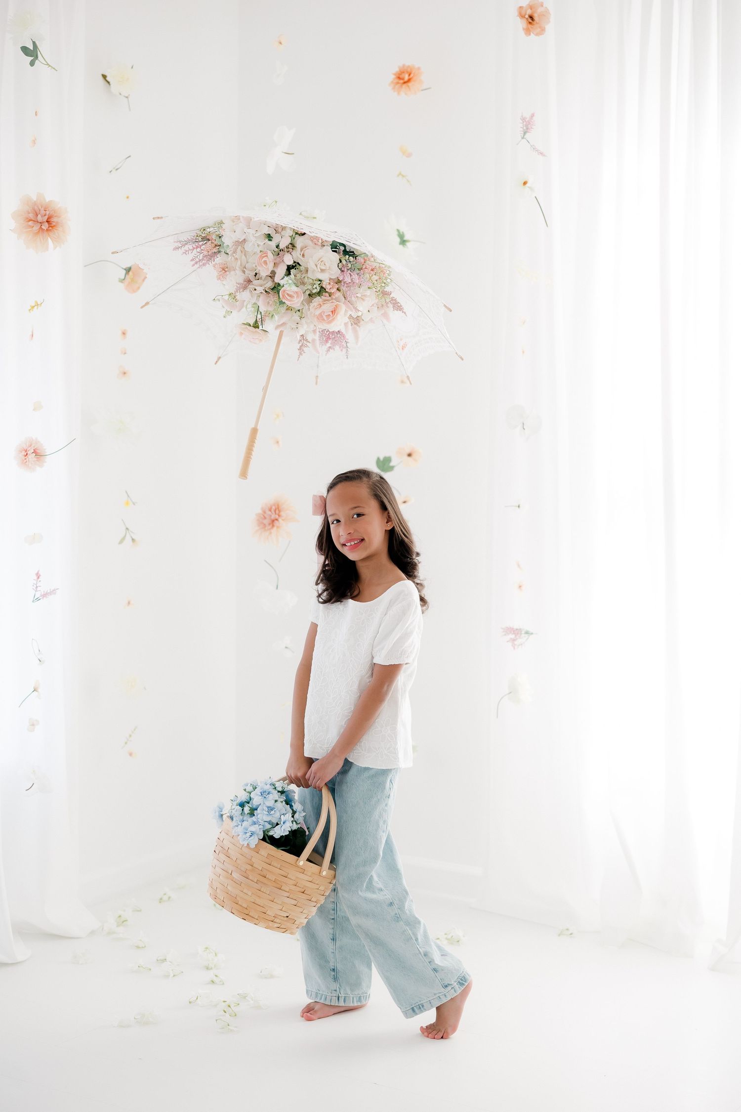 A person in light blue pants and white top poses playfully in a bright white studio with delicate floral decorations.