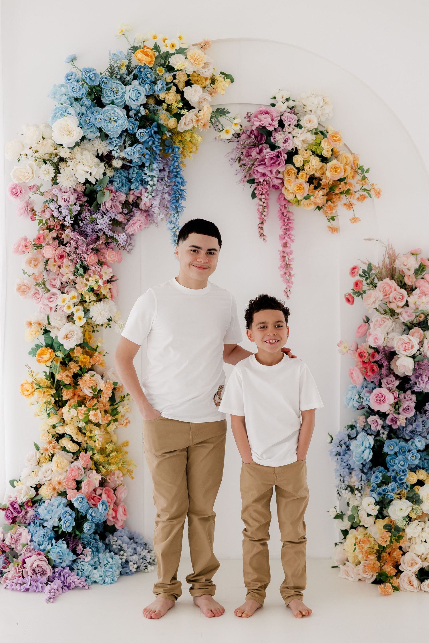 Two people in matching white shirts and khaki pants pose against a colorful floral backdrop in a photo series.