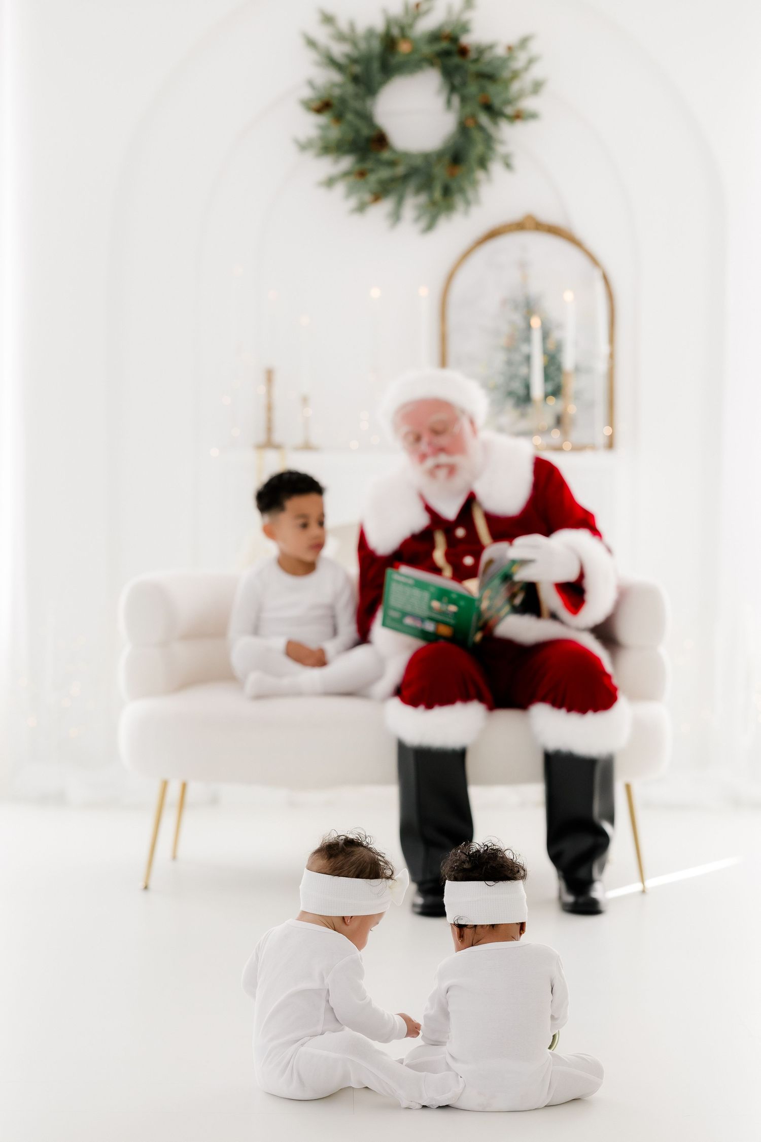 Santa Claus reads a book to children in white pajamas in a bright, festive white room with holiday decor.