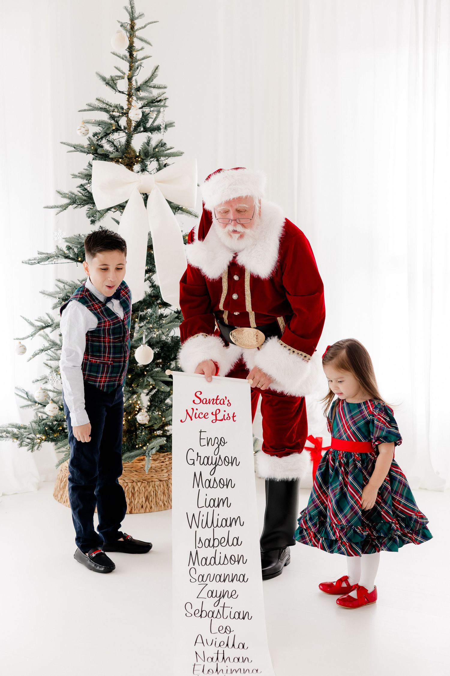 Santa Claus holds his Nice List scroll with two children beside a decorated Christmas tree in a bright white room.