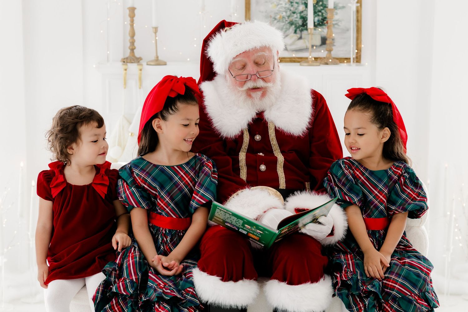 Santa Claus reading a Christmas book with three girls in festive plaid dresses and red bows.