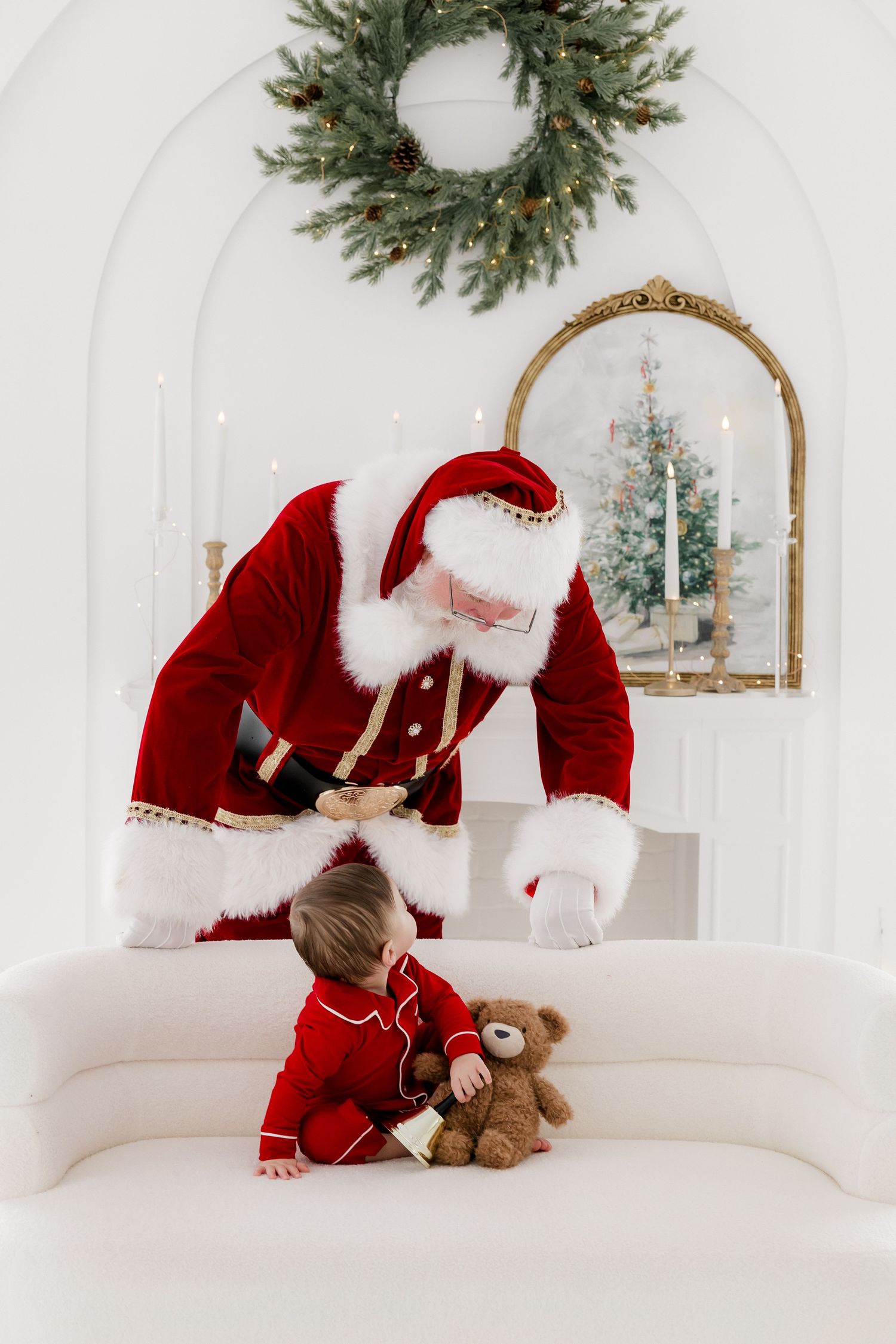 Santa Claus leaning toward a toddler in red pajamas holding a teddy bear on a white sofa with Christmas wreath decor.