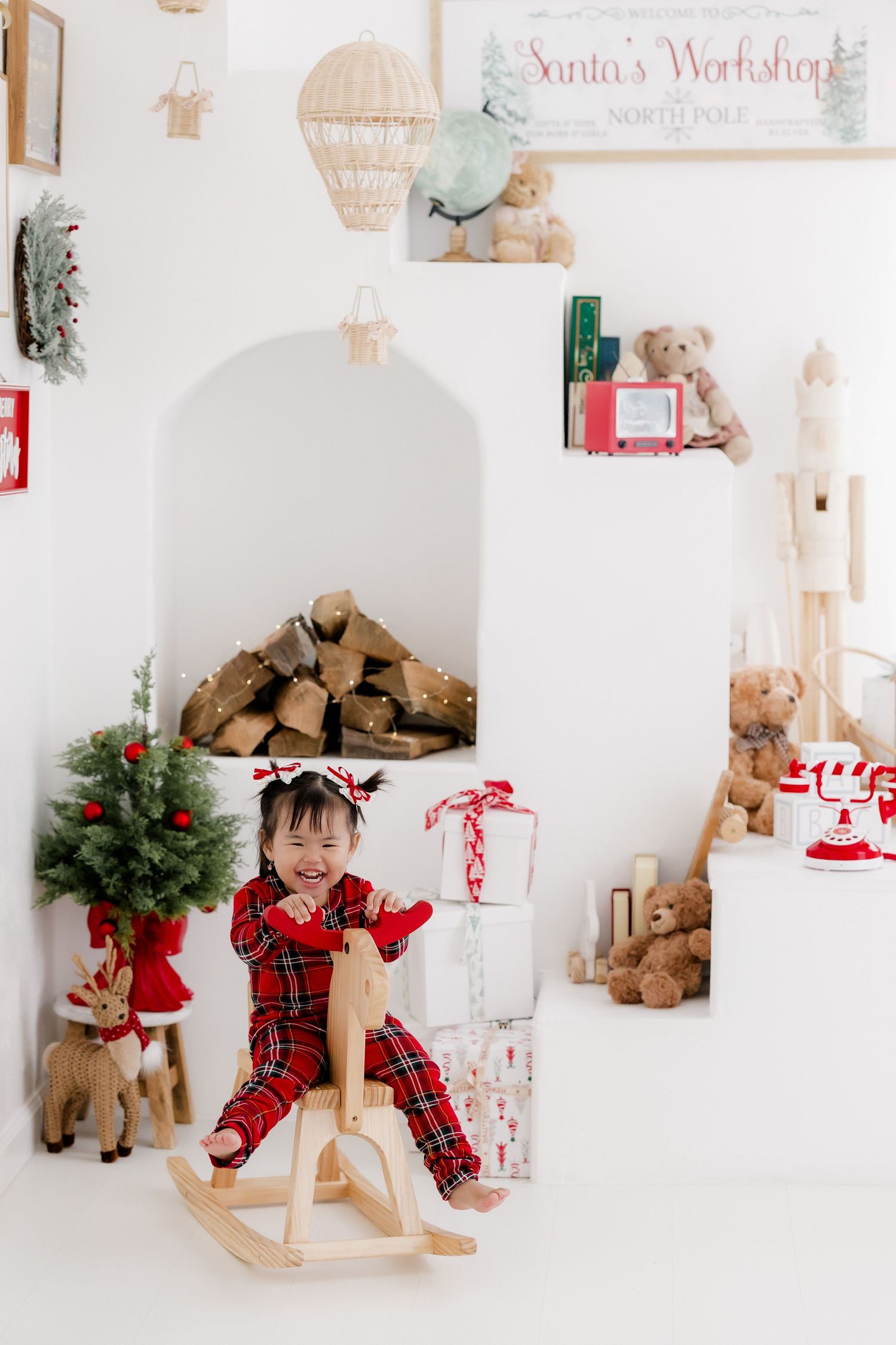 Young girl in red plaid pajamas rides wooden rocking horse in festive Christmas room with Santa's Workshop decor.