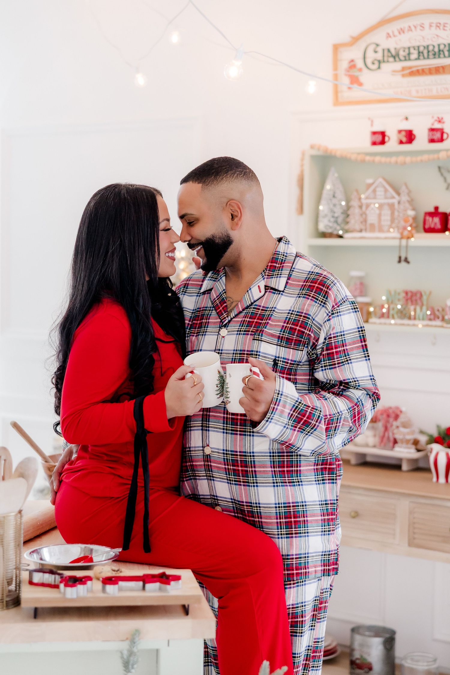 Couple in matching Christmas pajamas sharing mugs in a festive holiday kitchen decorated with gingerbread house.