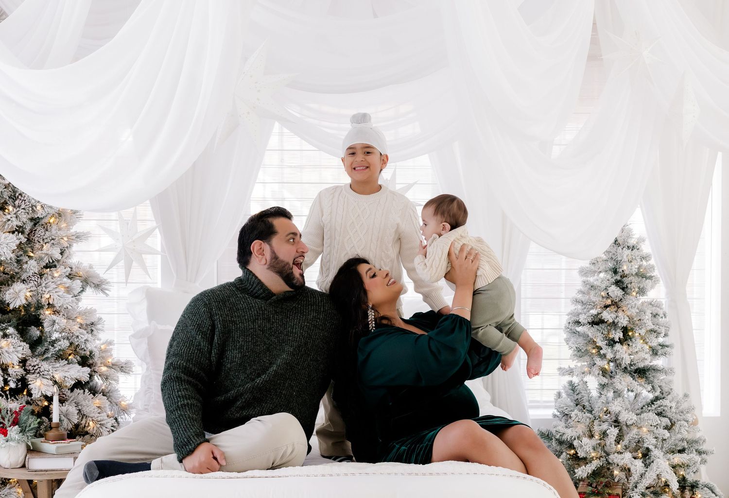 Family posing for a festive Christmas photo surrounded by snowy white trees and draped white fabric backdrop.