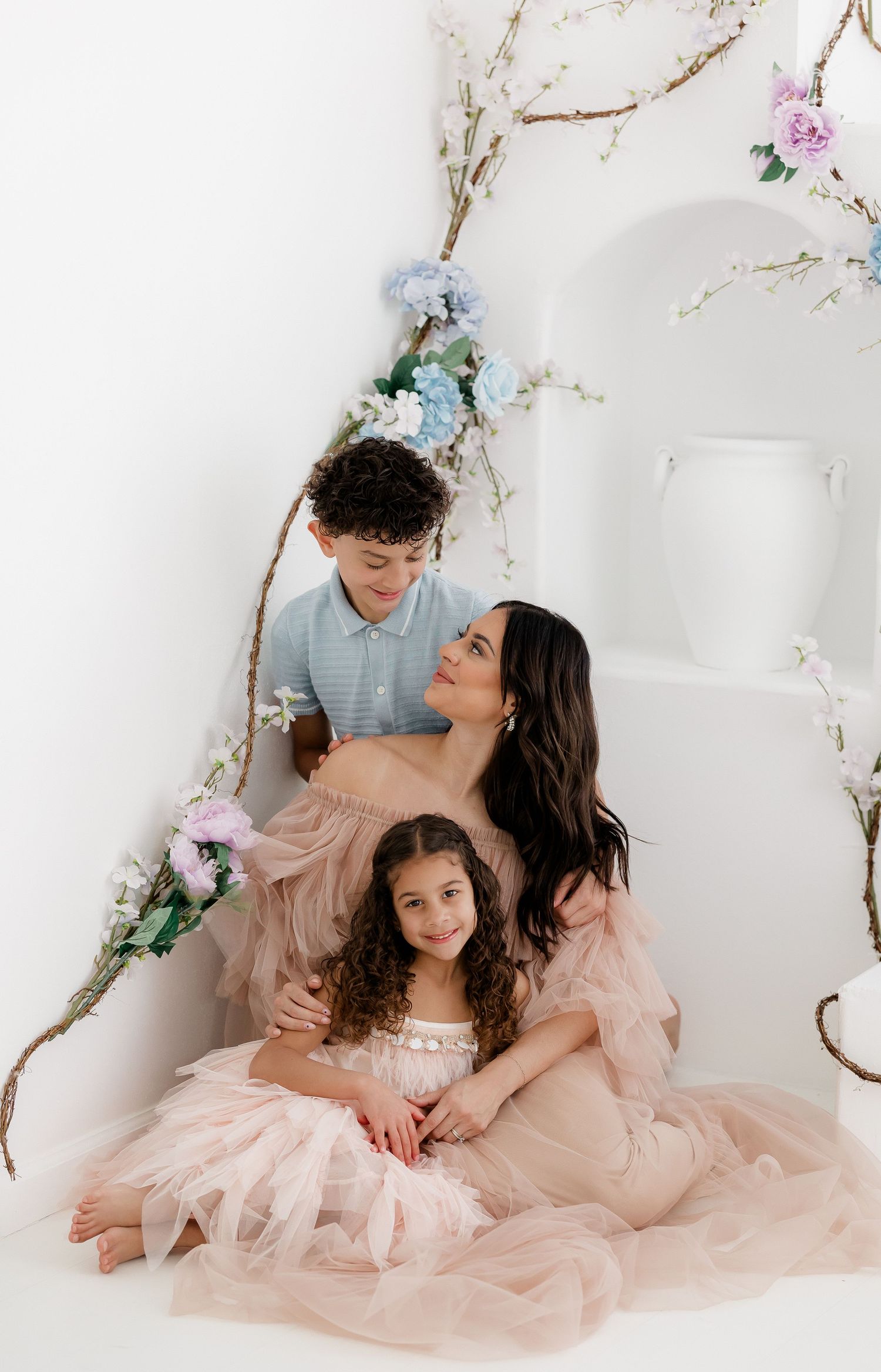 A family in matching blush pink tulle dresses poses together against a white backdrop with delicate floral decorations.