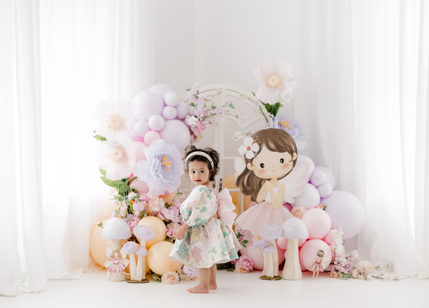 Toddler girl in floral dress poses with pastel balloons, paper flowers, and a fairy tale doll cutout backdrop.