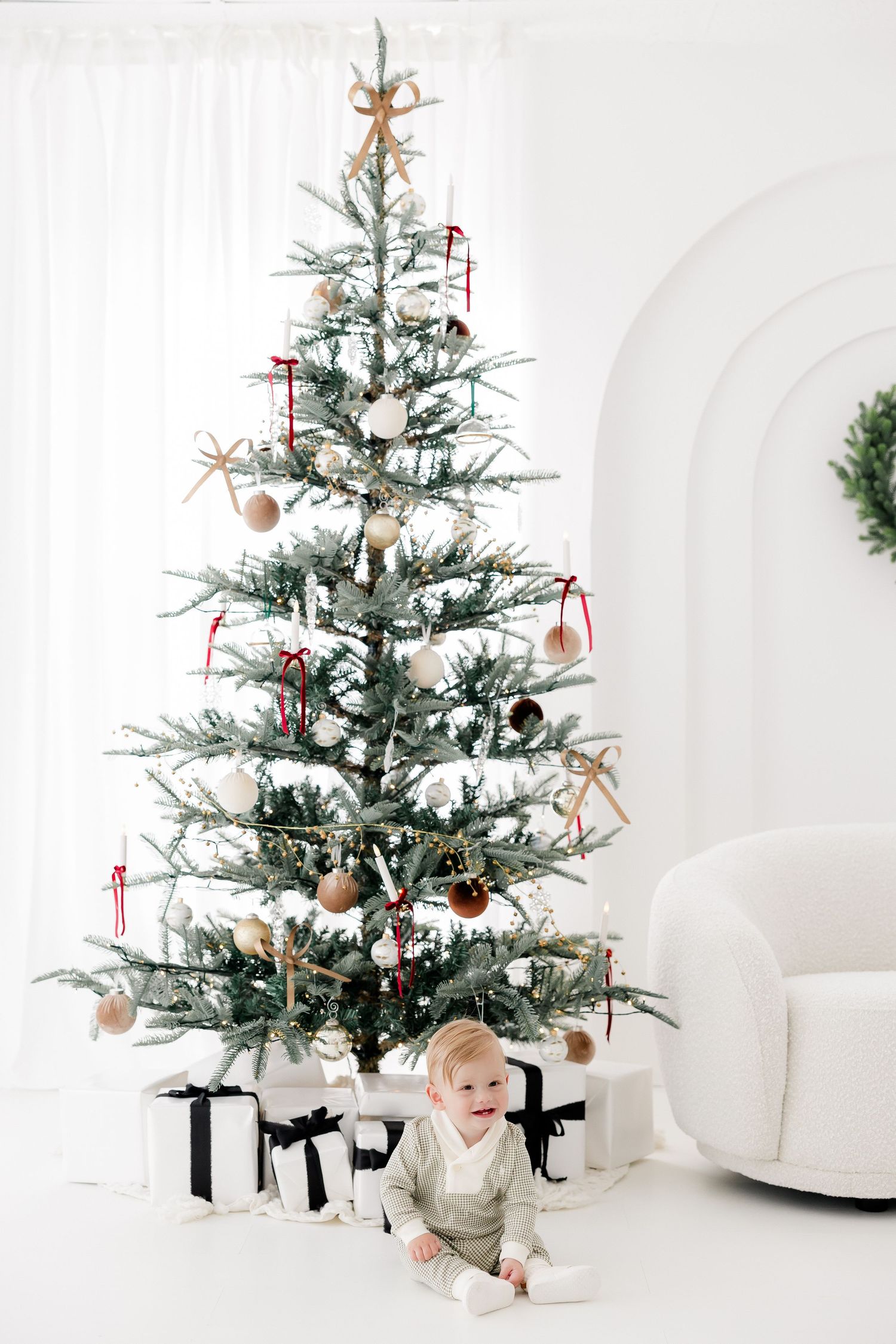 Child explores presents under decorated Christmas tree in bright white studio setting.