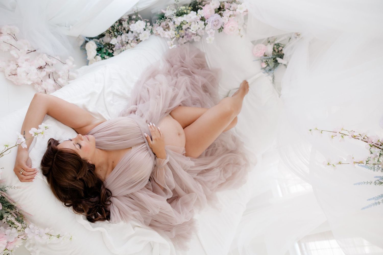 Romantic boudoir photoshoot featuring soft pink tulle fabric and delicate floral arrangements in ethereal lighting.