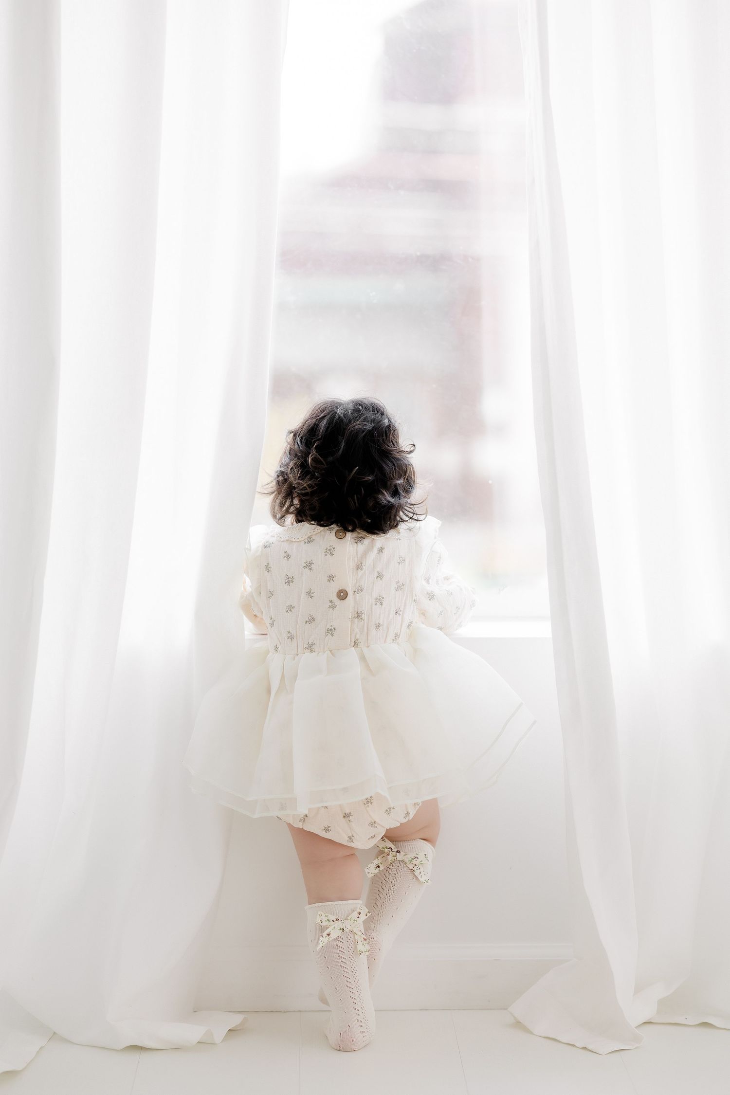 Toddler girl in white dress and lace socks stands on tiptoes gazing out a bright window with sheer curtains.