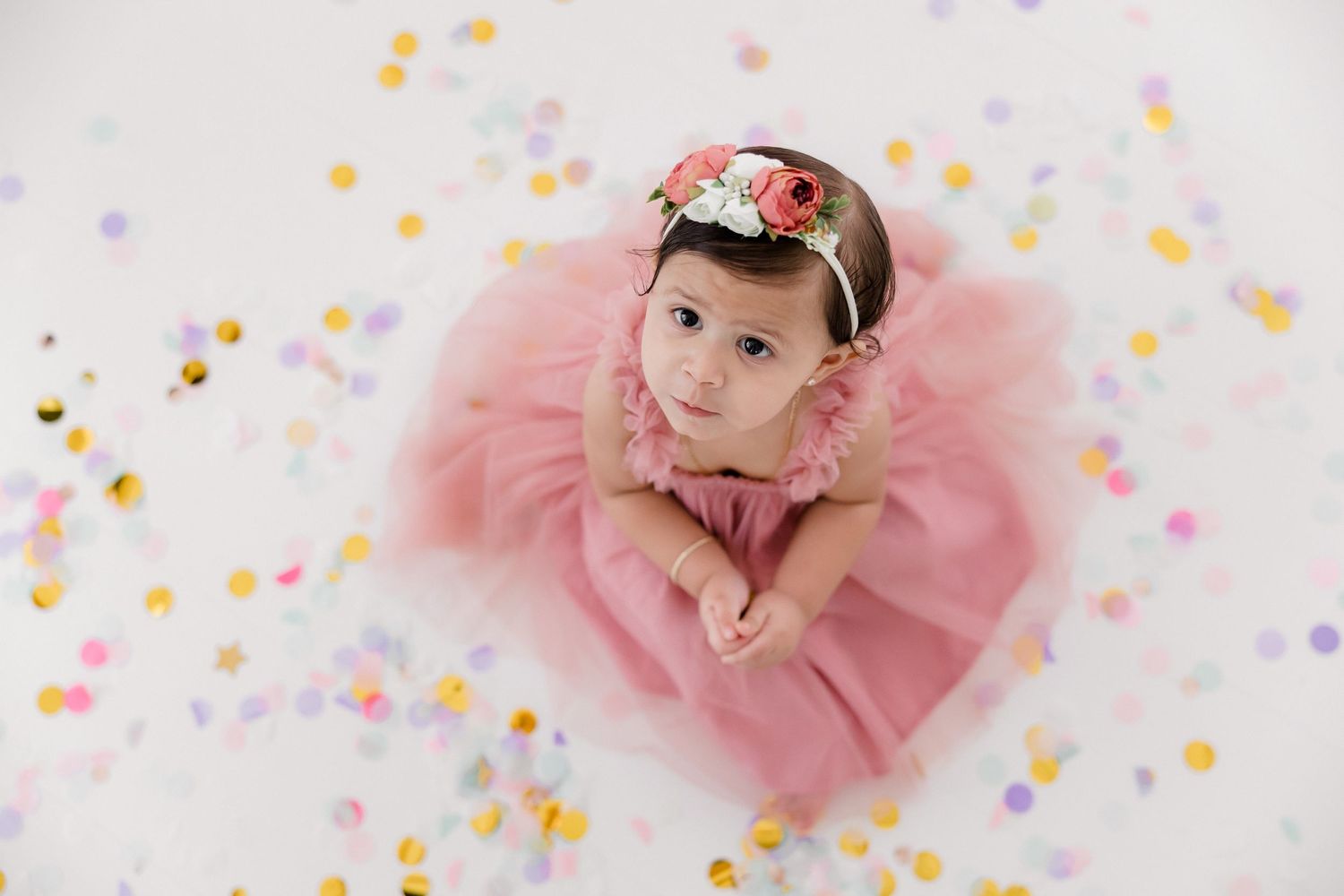 A baby in a pink tutu dress lies on a white backdrop surrounded by colorful confetti during a photo session.
