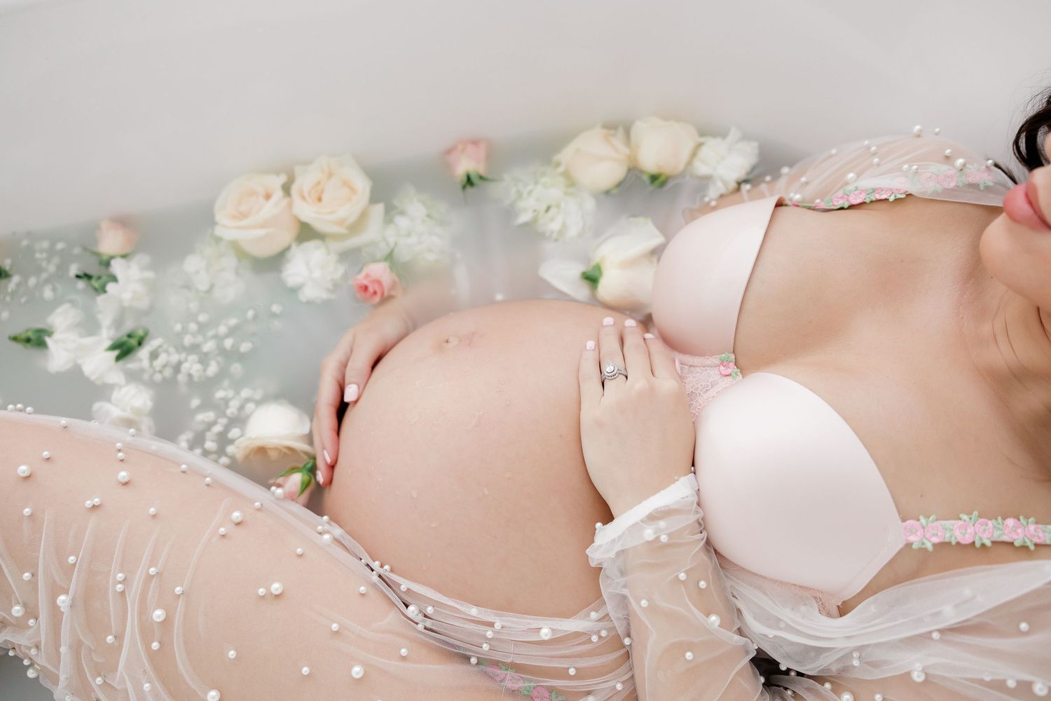 A pregnant woman in white lingerie poses among delicate flowers and lace decorations in a milk bath during a maternity photoshoot.