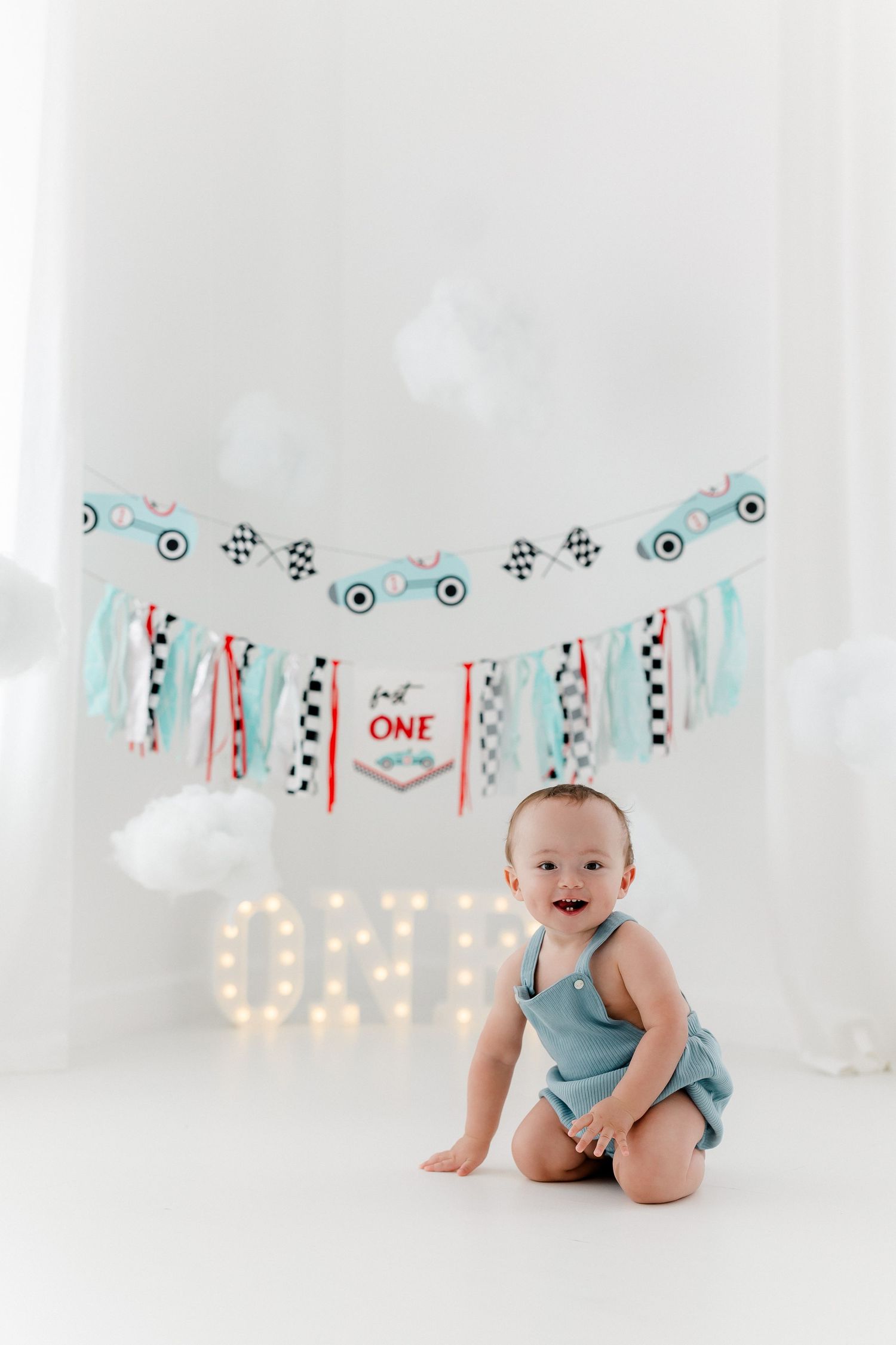 Baby in blue romper sits on white backdrop with festive birthday banner and lights for first birthday photo session.